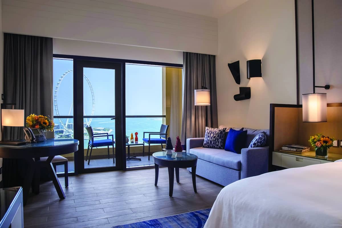 A hotel room with a bed, a sofa, a dining table, and a balcony with a view of a Ferris wheel.