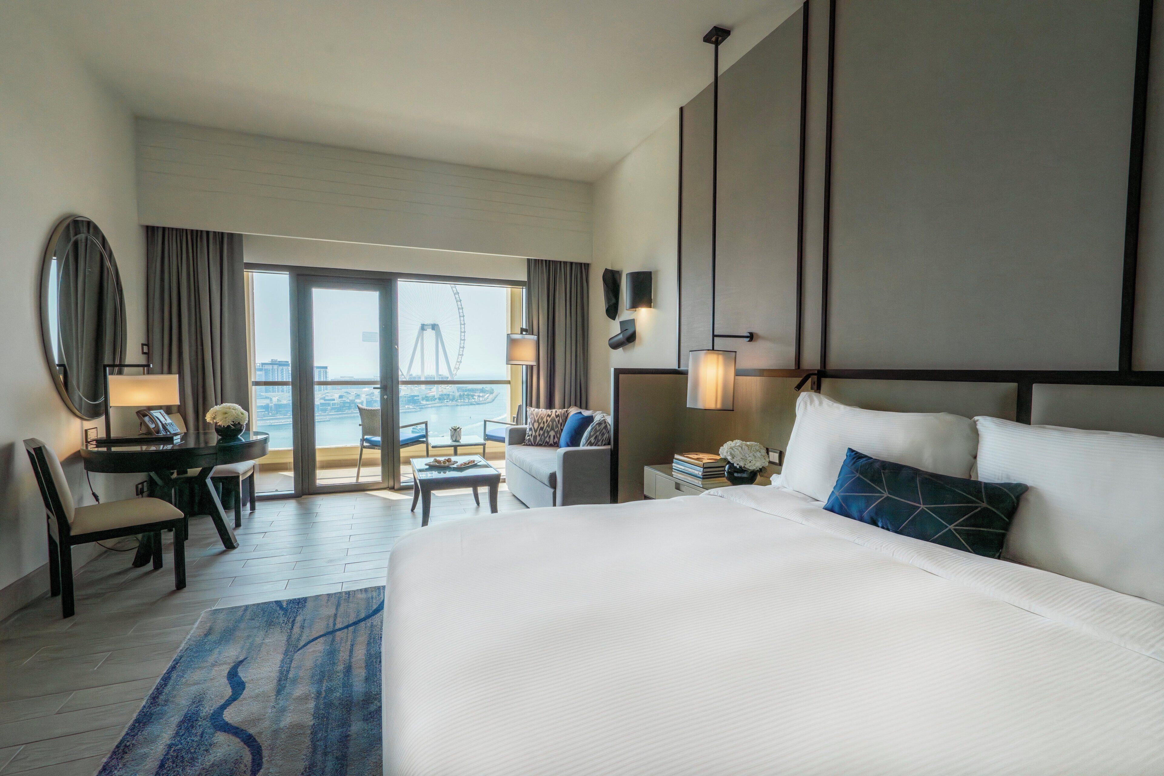 A hotel room with a large bed, a desk with a chair, a small table, a sofa, and a view of a Ferris wheel.