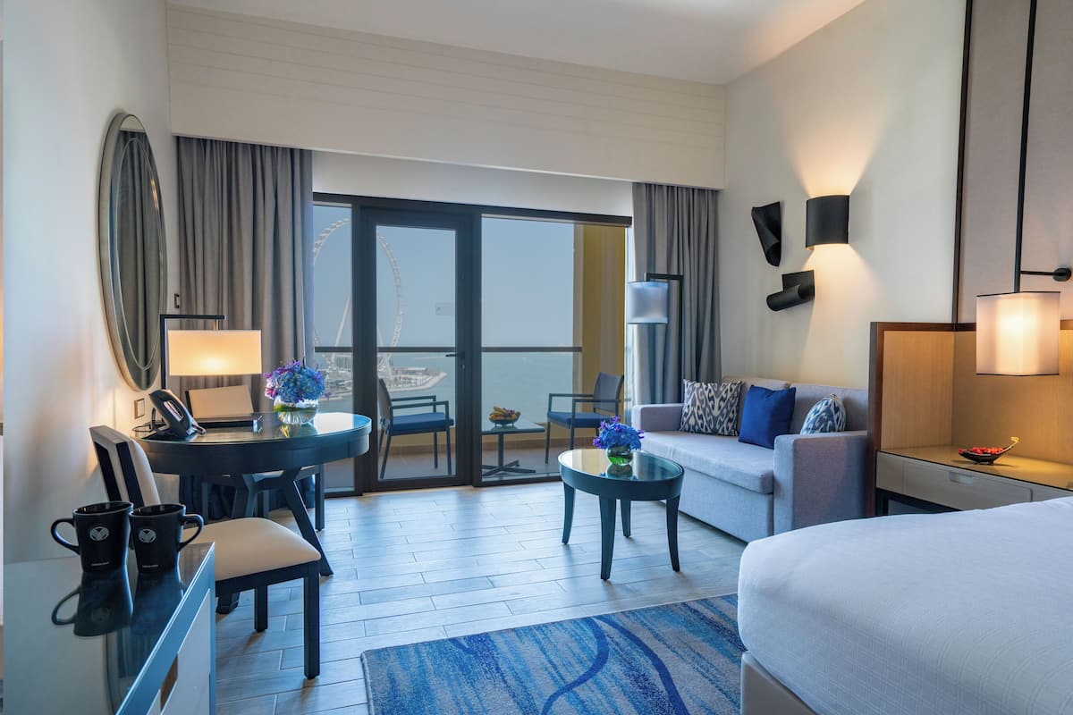 A hotel room with a bed, a sofa, a dining table with chairs, a desk with a chair, a small table with a vase of flowers, and a large window with curtains.