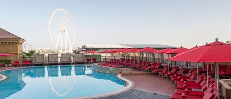 A swimming pool with red lounge chairs and umbrellas, a Ferris wheel in the background, and a clear sky.