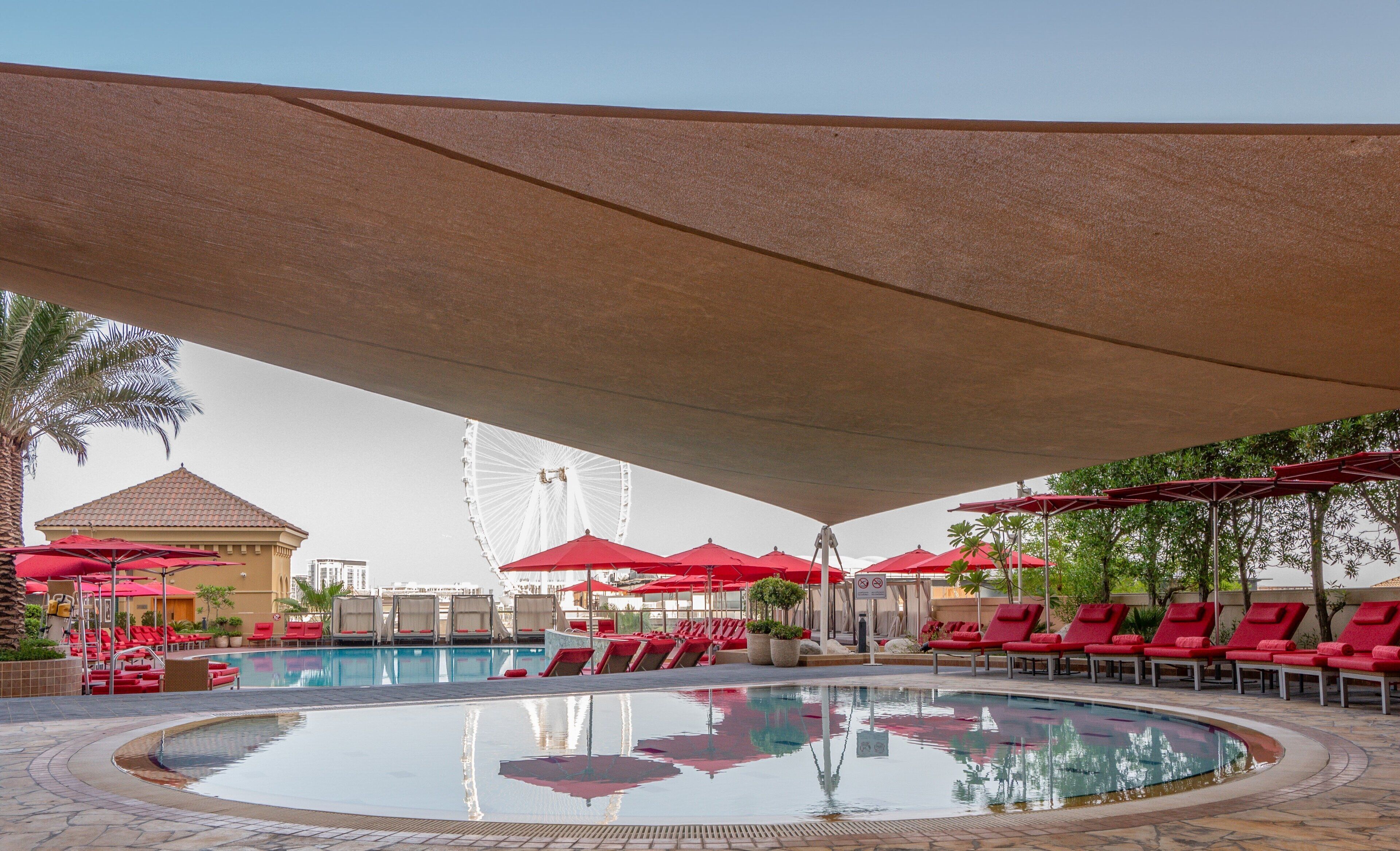 A swimming pool with red umbrellas and lounge chairs, a Ferris wheel in the background, and a palm tree.