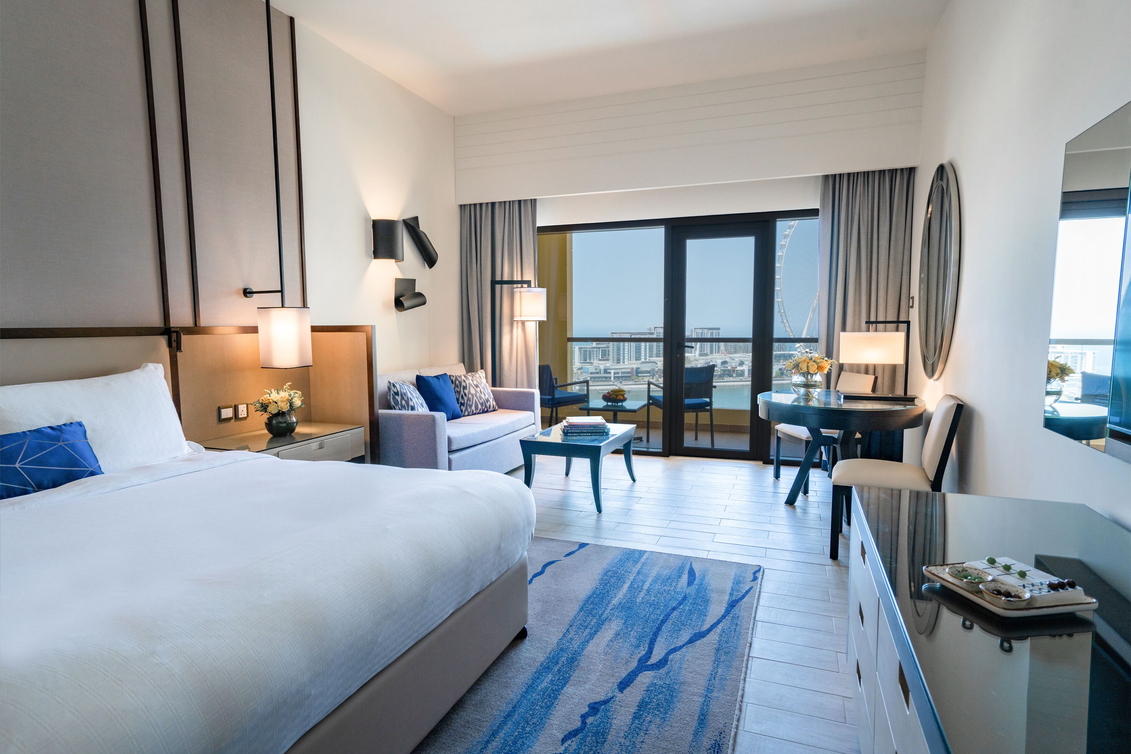 A hotel room with a large bed, a sofa, a desk, and a balcony with a view of the city.