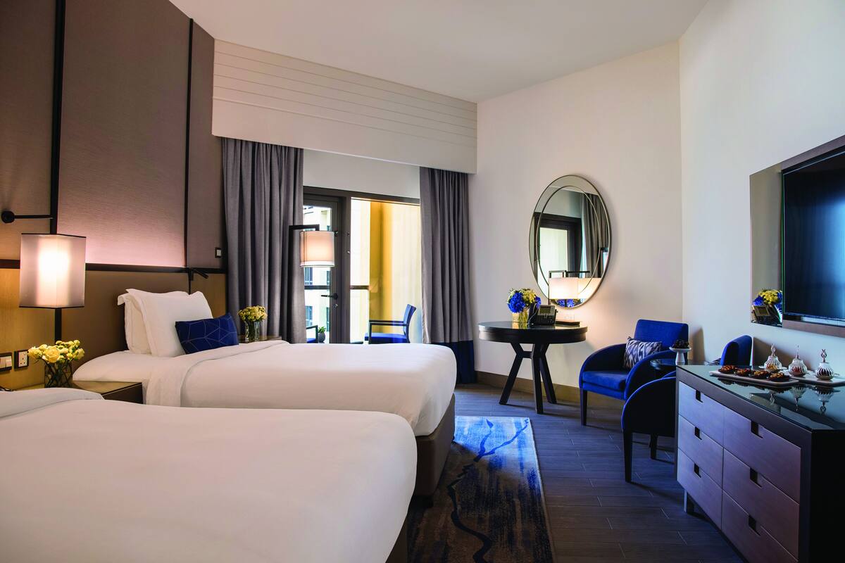A hotel room with two beds, a desk, a chair, a dresser, a mirror, and a TV.