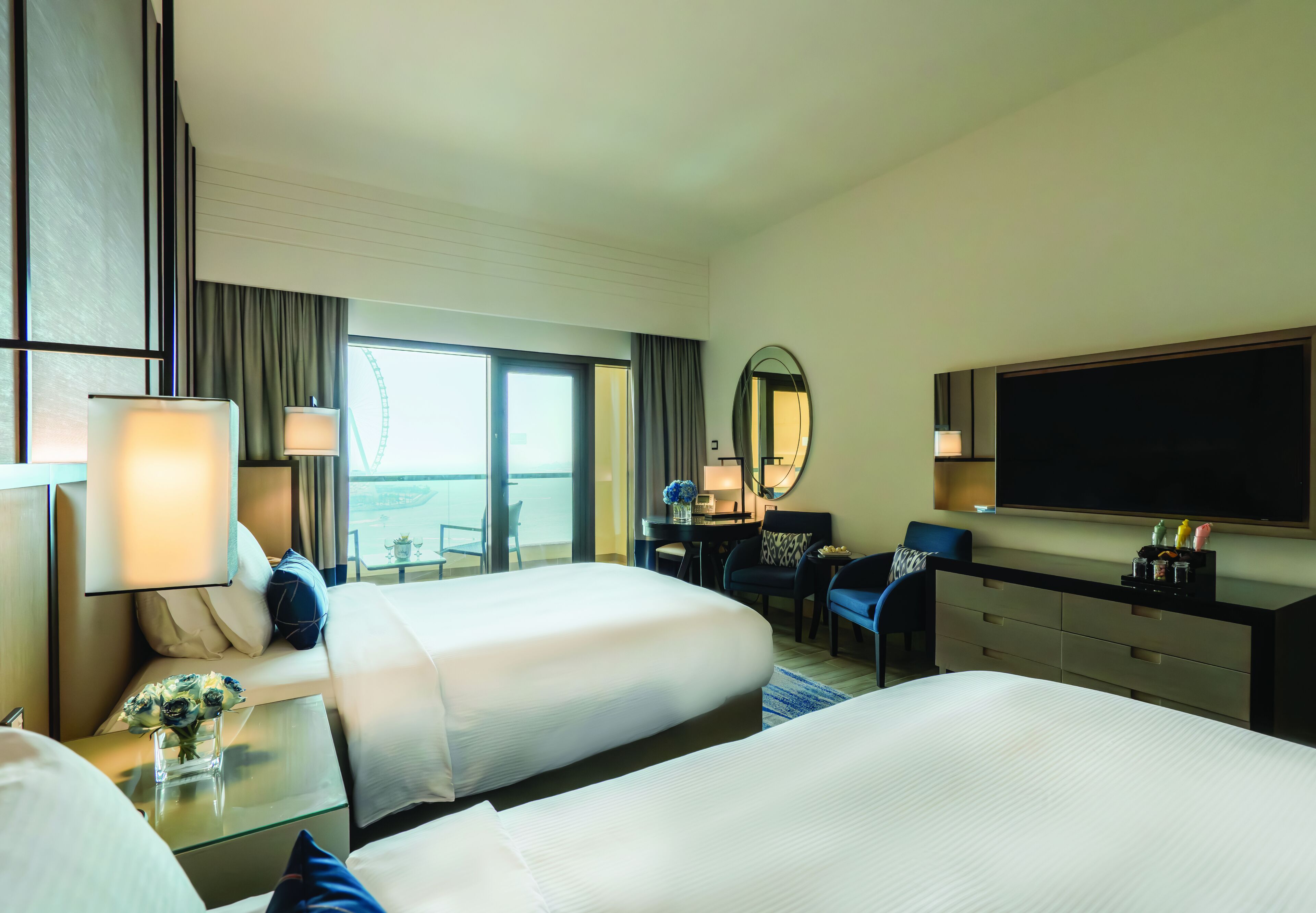 A hotel room with two beds, a desk, a chair, a TV, and a large window.