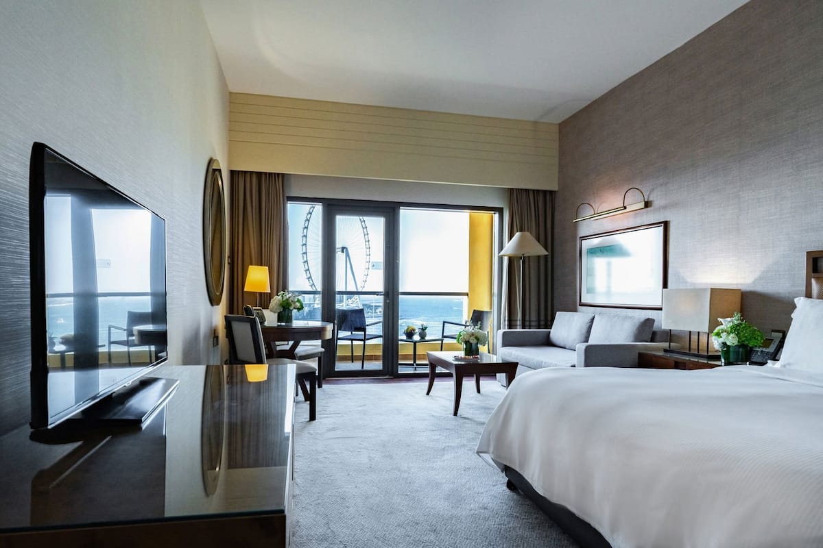 A hotel room with a bed, a desk, a TV, a chair, a table, and a view of the ocean and a Ferris wheel.