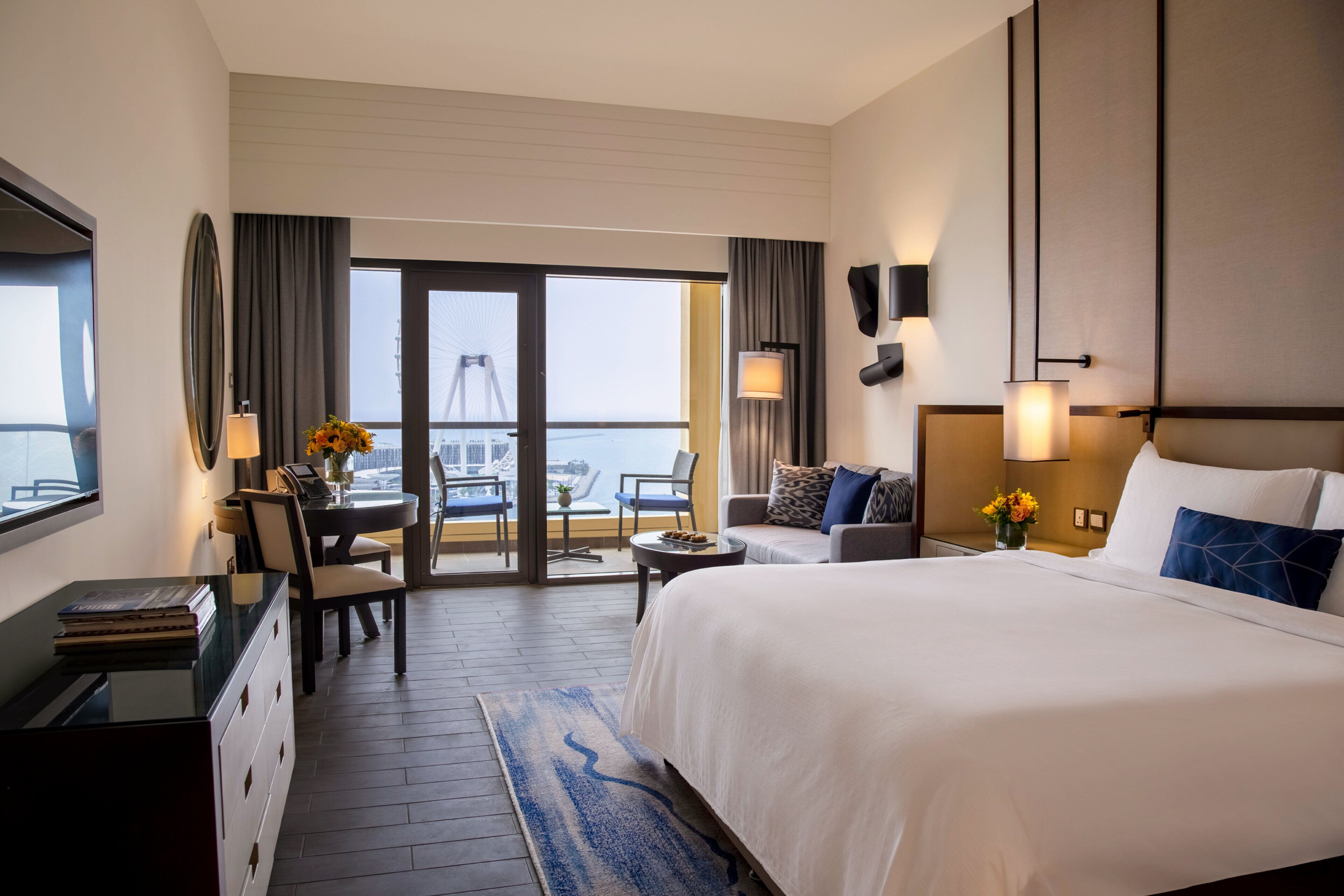 A hotel room with a large bed, a desk, a chair, a dining table, and a view of a Ferris wheel through a glass door.