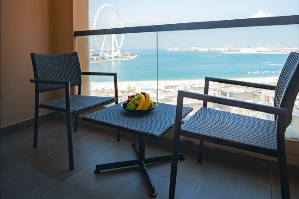 A balcony with two chairs and a table, a bowl of fruit, and a view of a Ferris wheel and coastline.