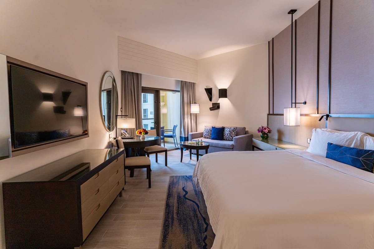 A modern hotel room with a large bed, a desk with a chair, a sofa, a TV, and a large mirror.