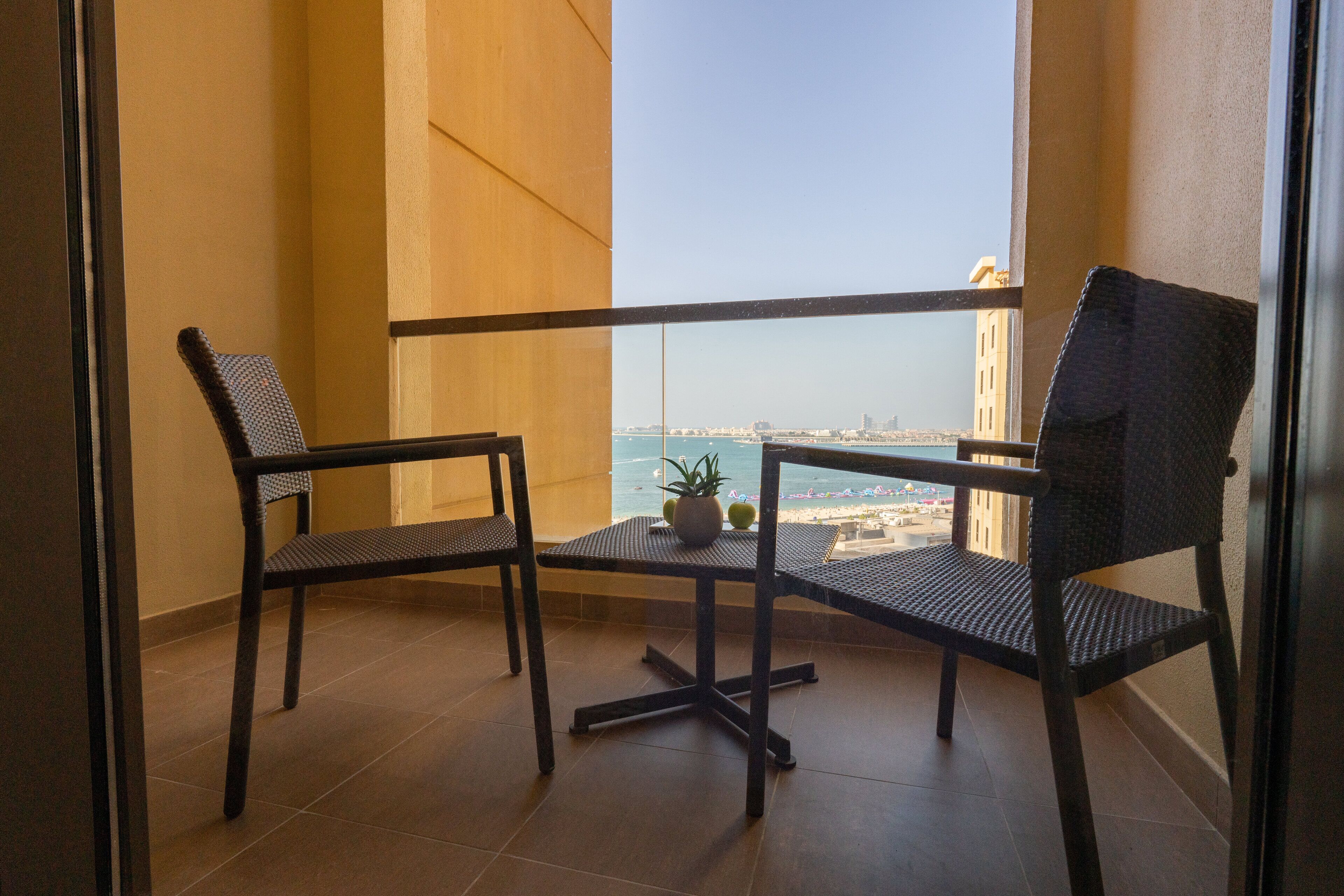 A balcony with two wicker chairs, a small table with a potted plant, and a view of the sea and buildings.