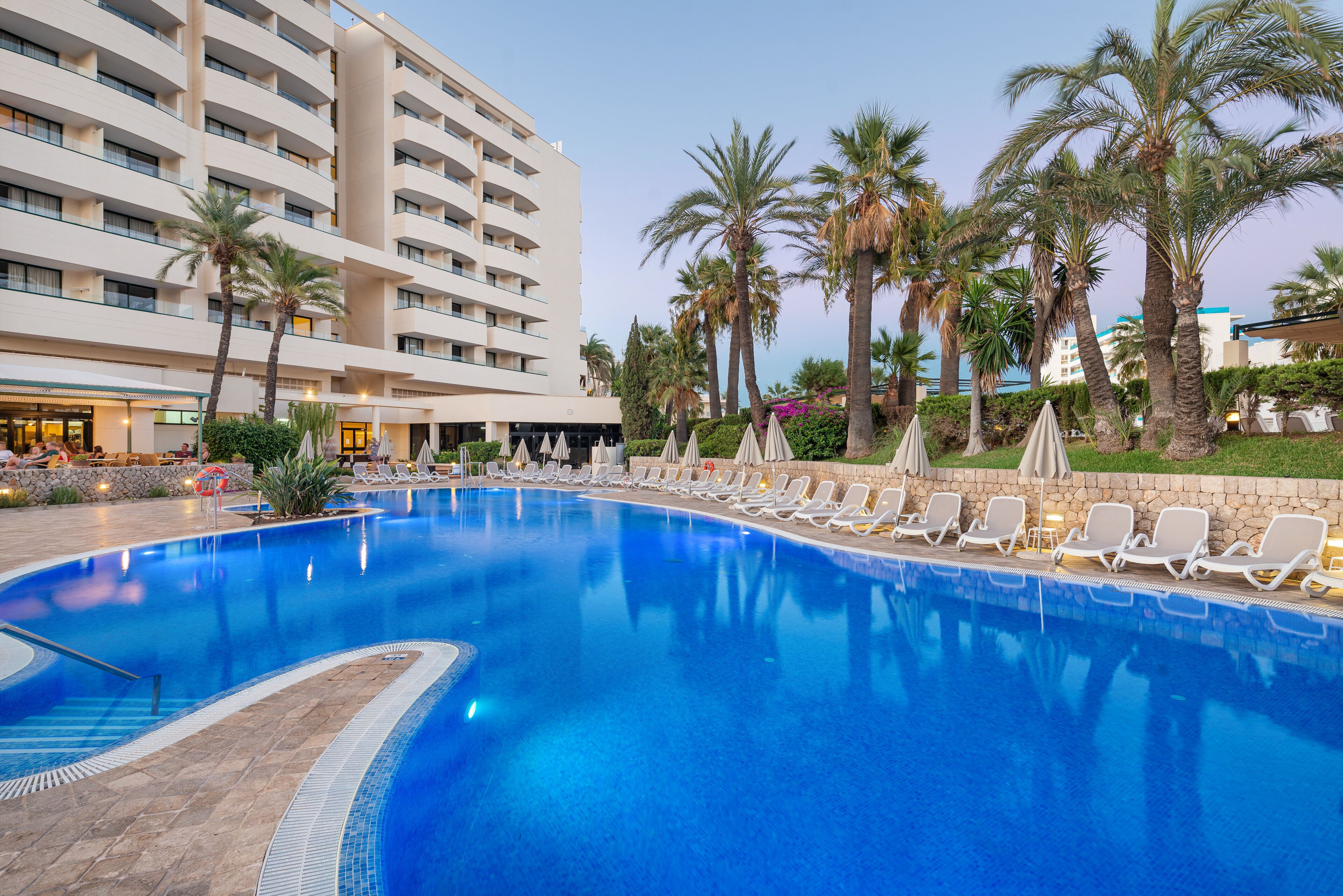 A hotel with a large swimming pool, palm trees, and sun loungers.