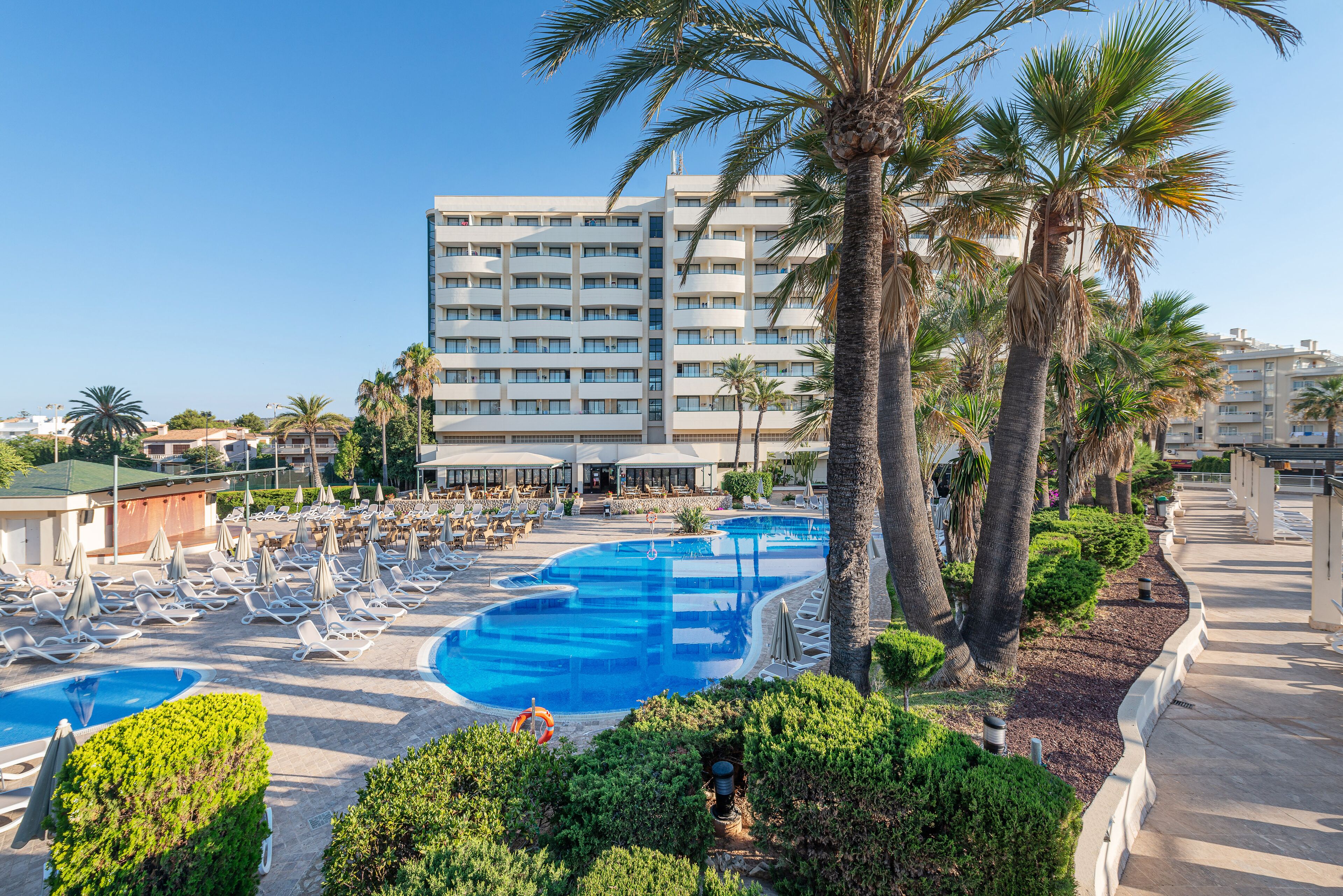Un hotel con un amplio espacio de piscina, numerosas tumbonas y palmeras.