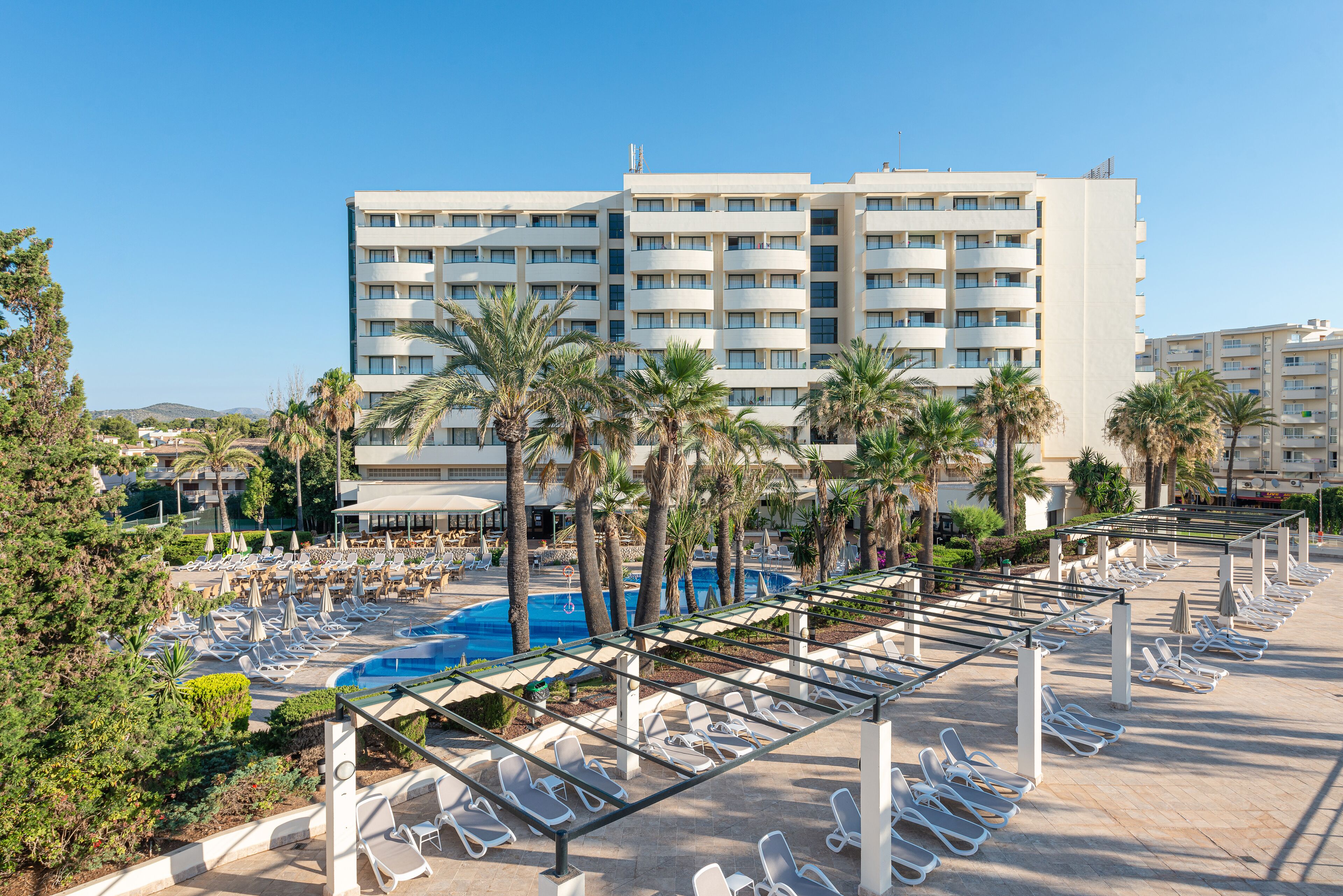 Un hotel con zona de piscina, palmeras y un cielo azul despejado.