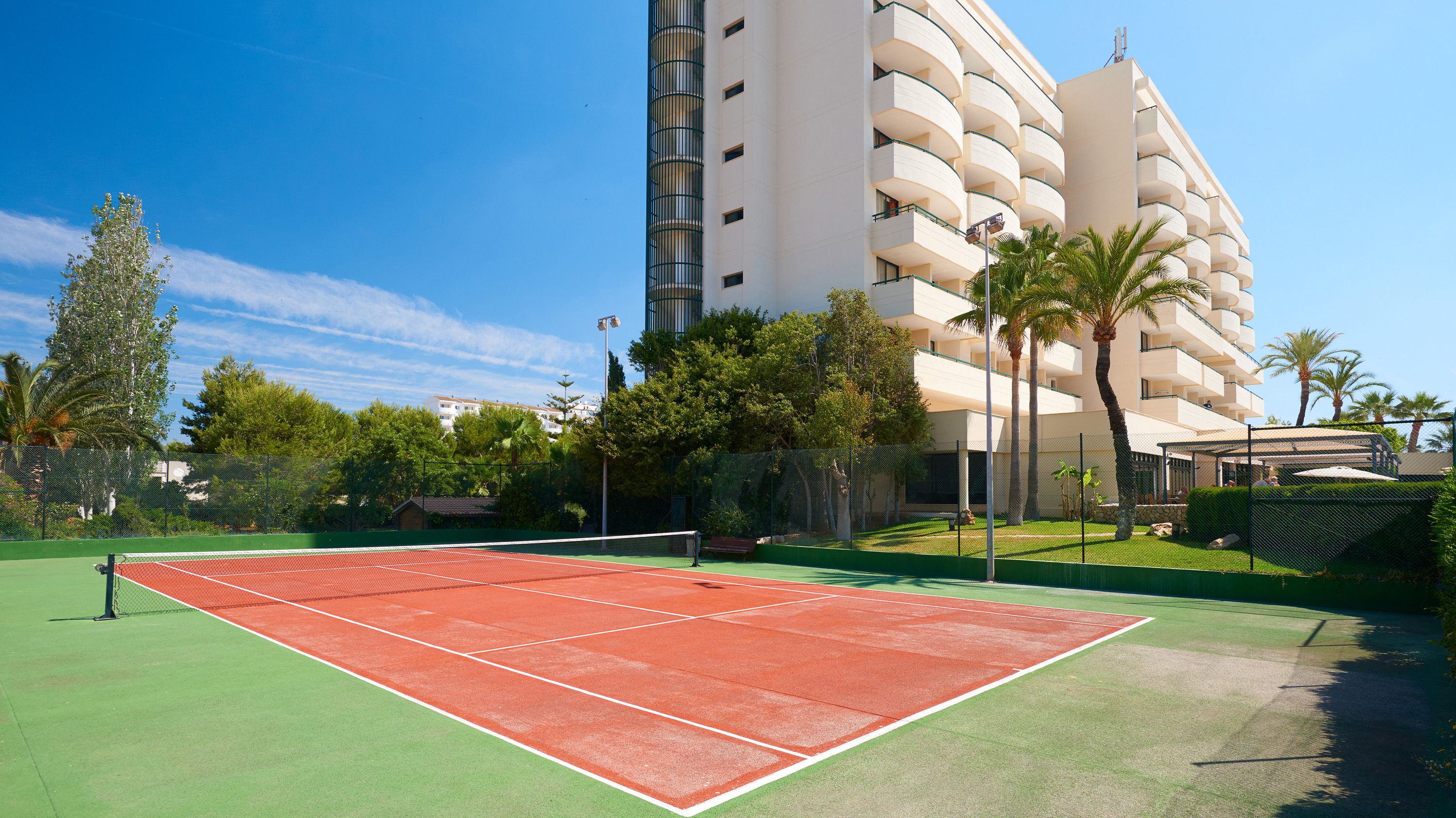 A tennis court with a net, surrounded by a green fence, and a tall building in the background.