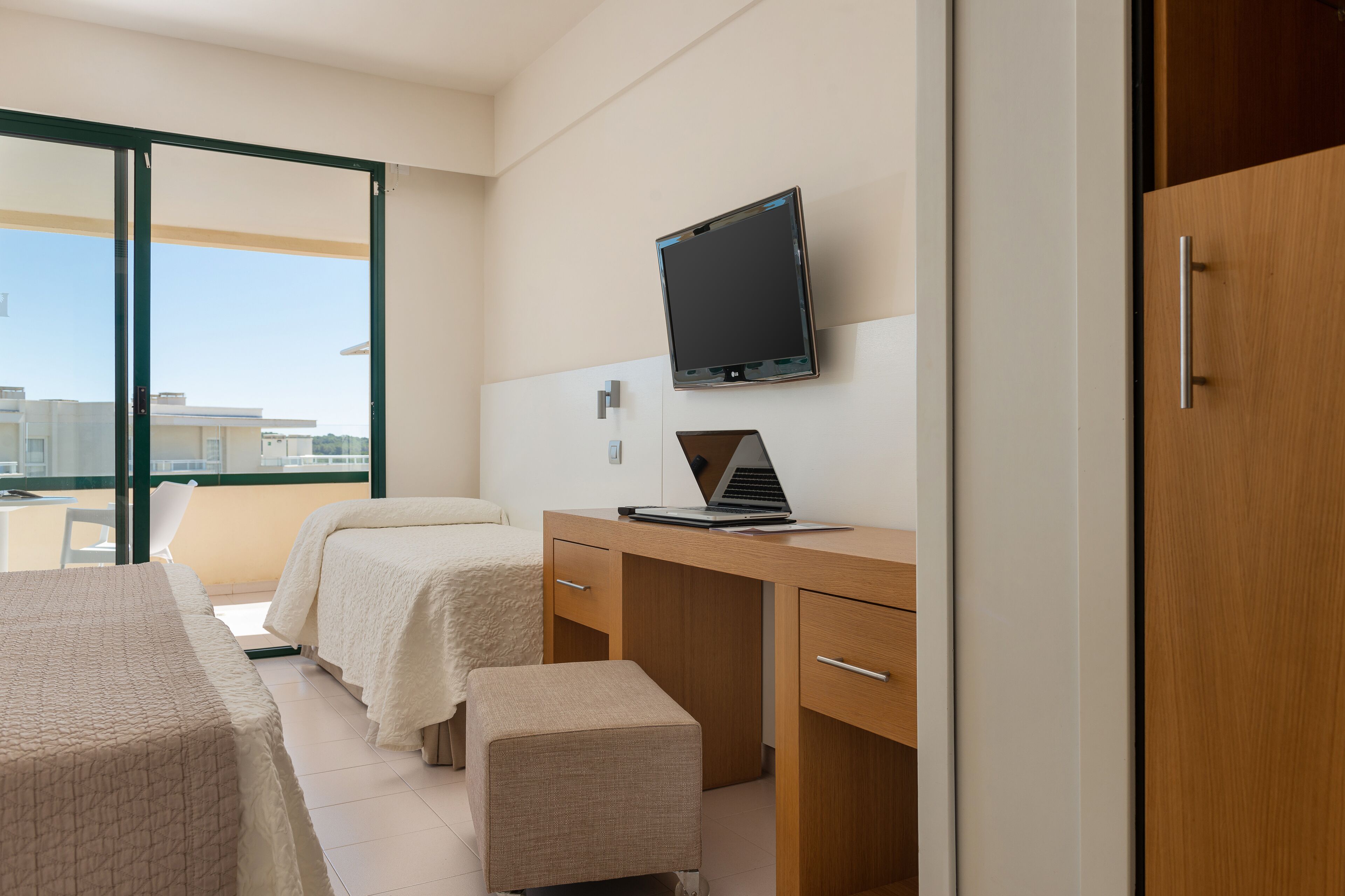 A hotel room with two beds, a desk with a laptop, a TV, and a sliding door to a balcony.
