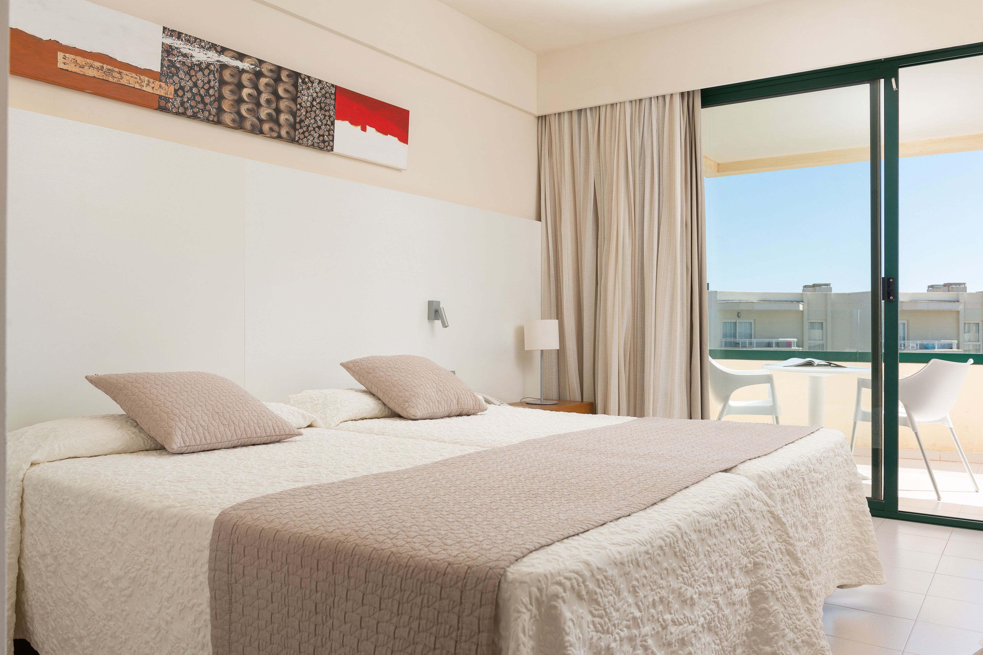 A hotel room with two beds, a large window with curtains, a view of buildings, and a table with chairs outside.