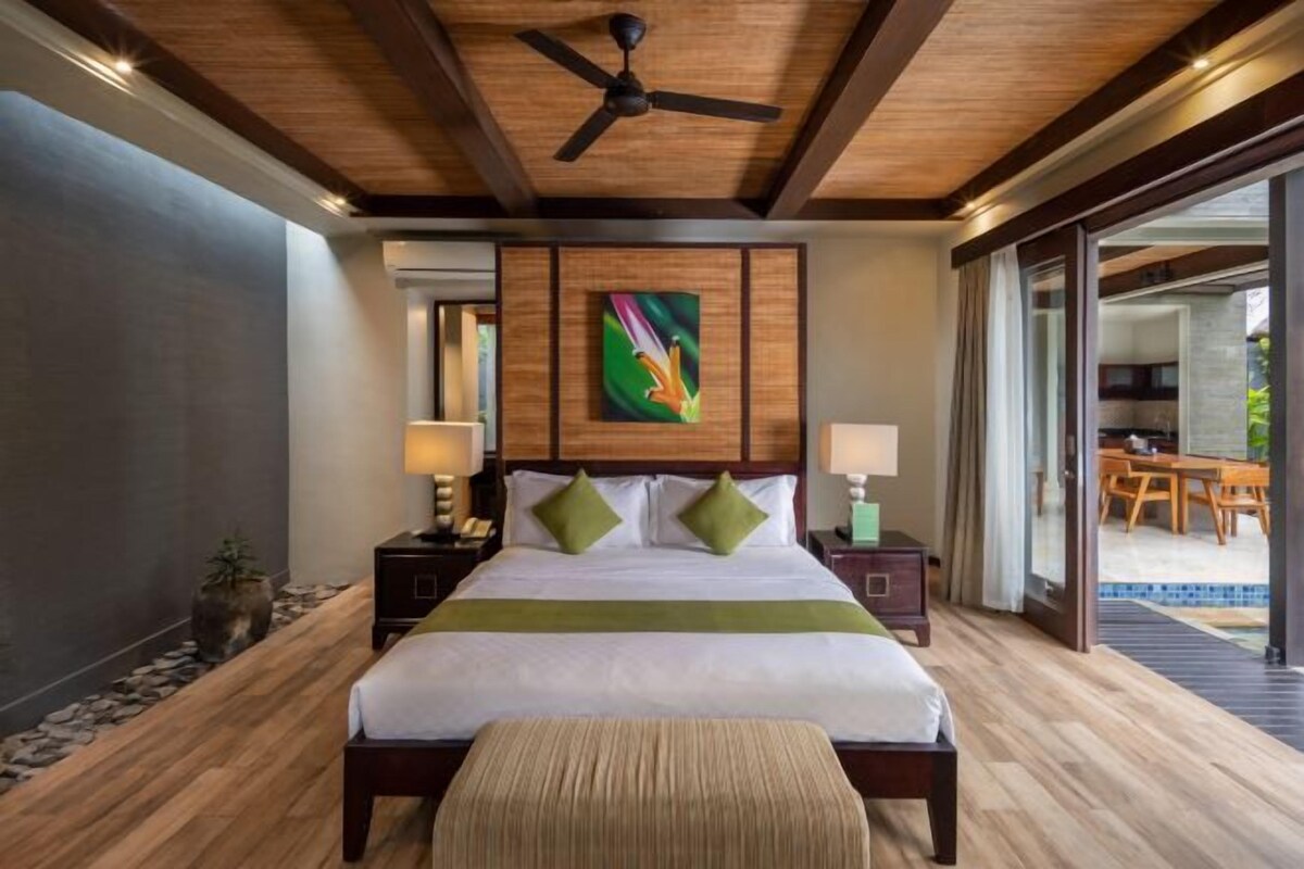 Habitación de hotel con una cama grande, techo de madera, ventilador de techo, un panel decorativo verde y vistas al patio exterior.