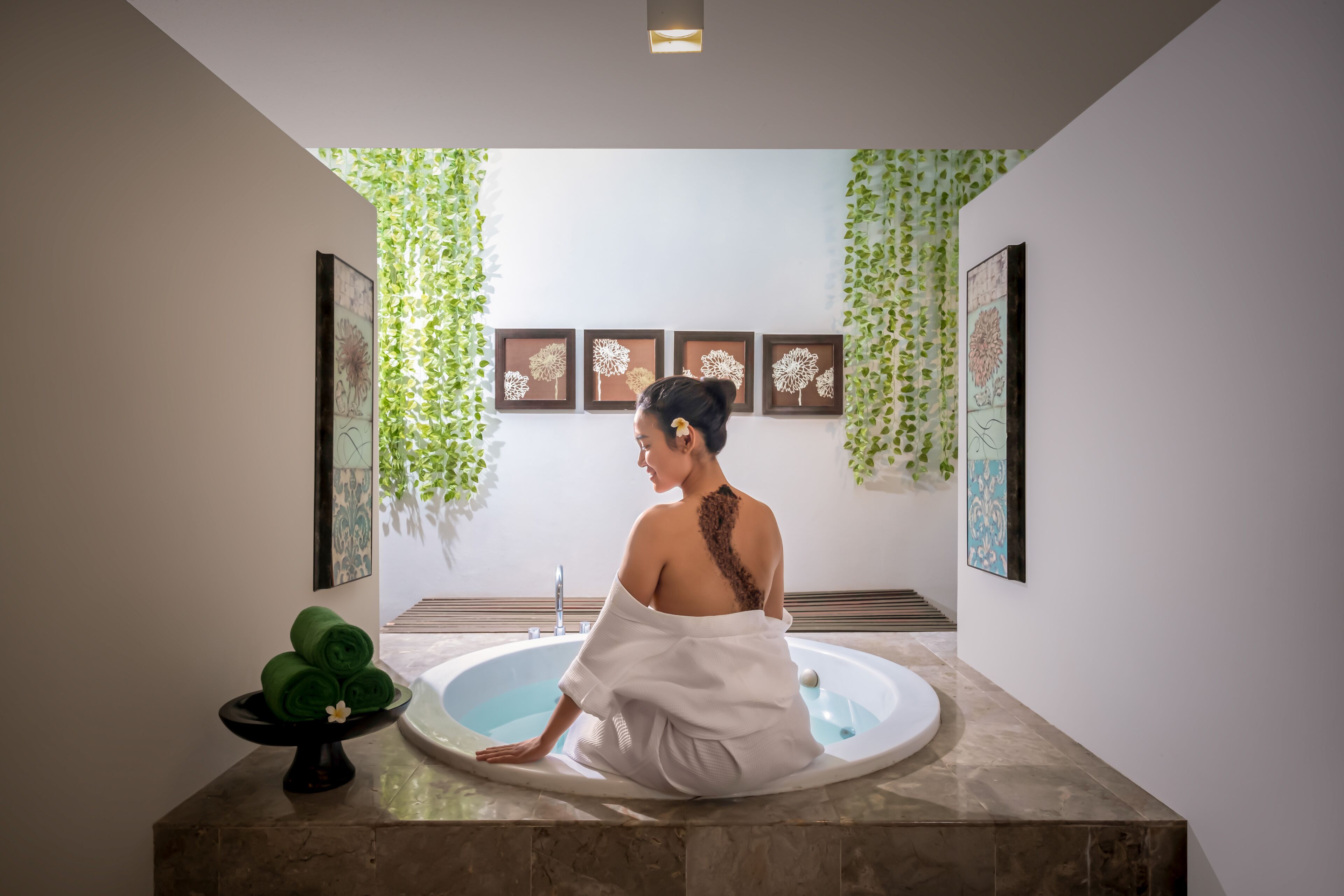 A person sitting in a bathtub in a spa-like setting with framed artwork and green foliage on the walls.