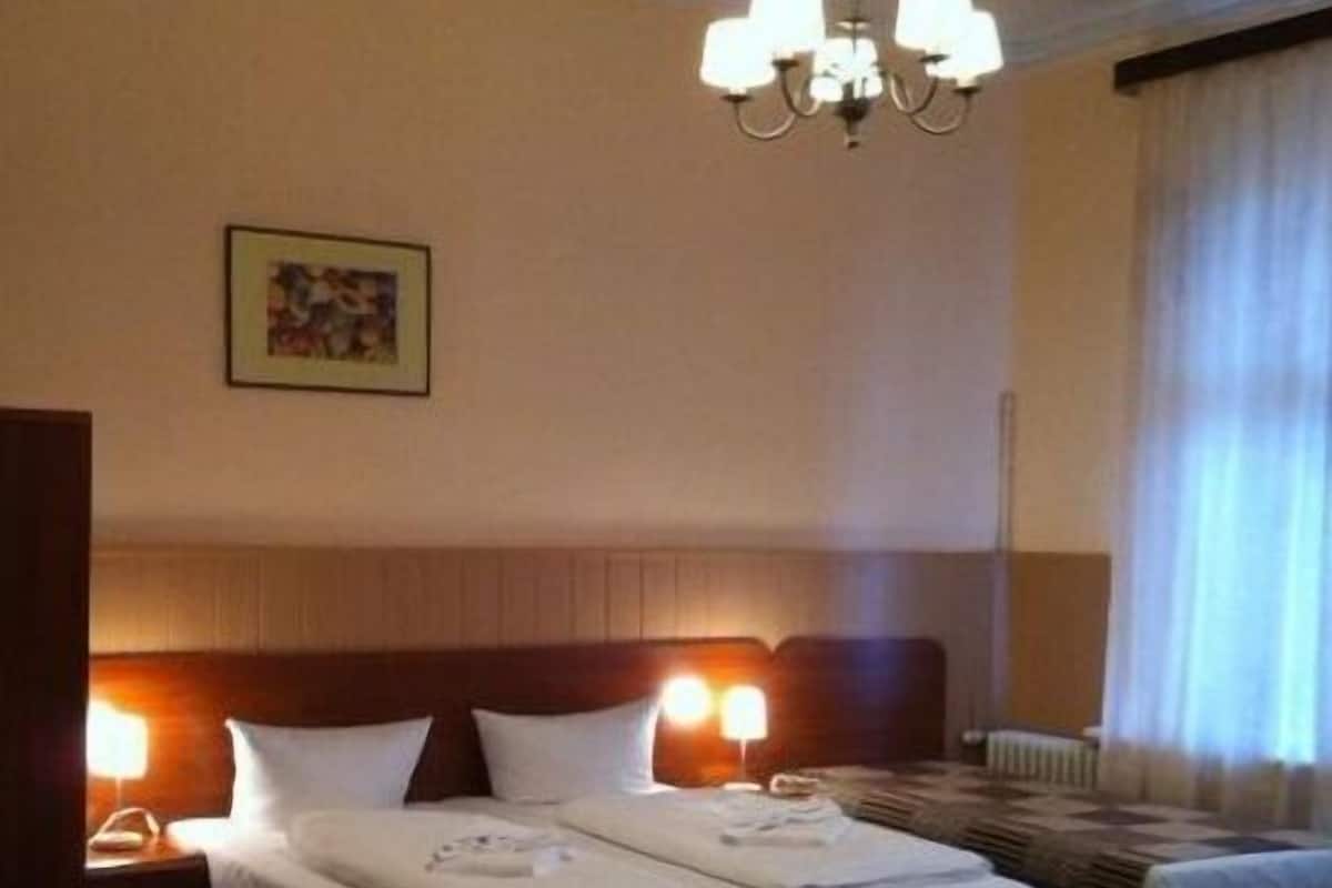 Une chambre d’hôtel avec un lit double, une table de chevet avec une lampe, un lustre et une fenêtre avec des rideaux.
