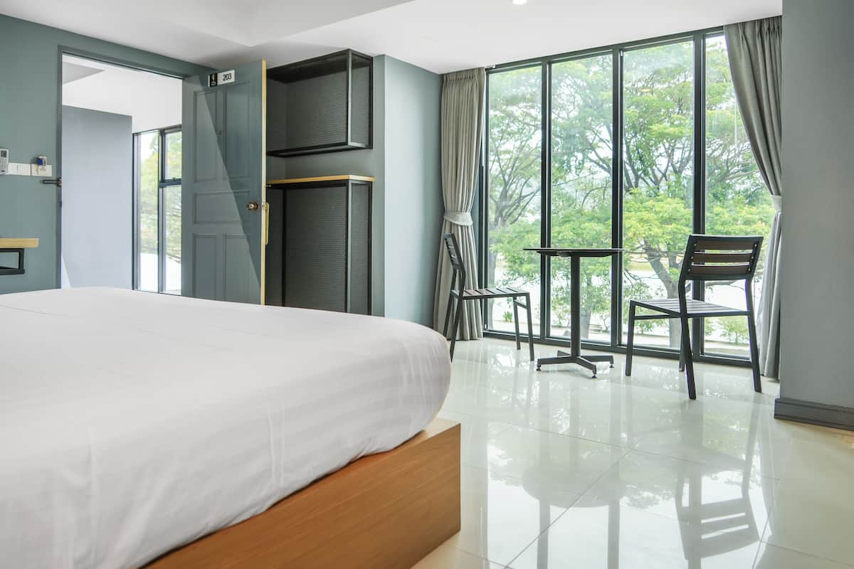A modern hotel room with a large bed, a balcony with a chair, and a view of trees.