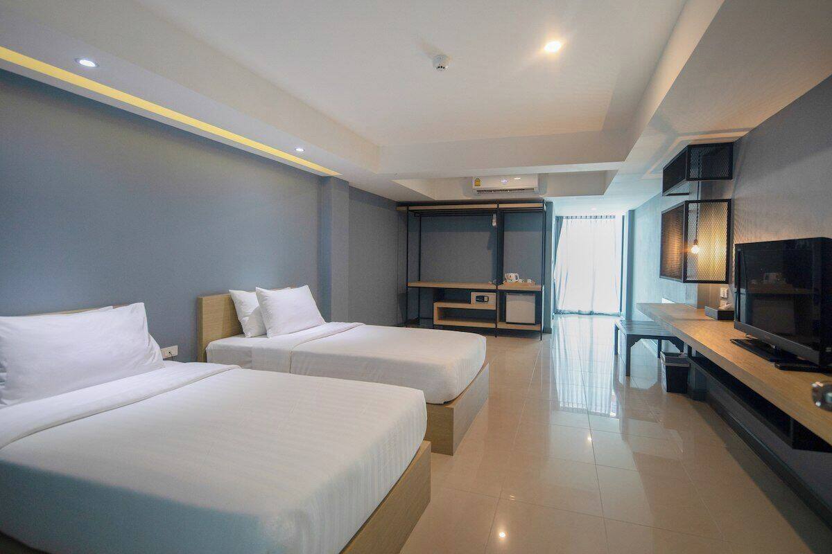 A modern hotel room with two single beds, a desk with a television, a wardrobe, and a seating area with a view of the street.