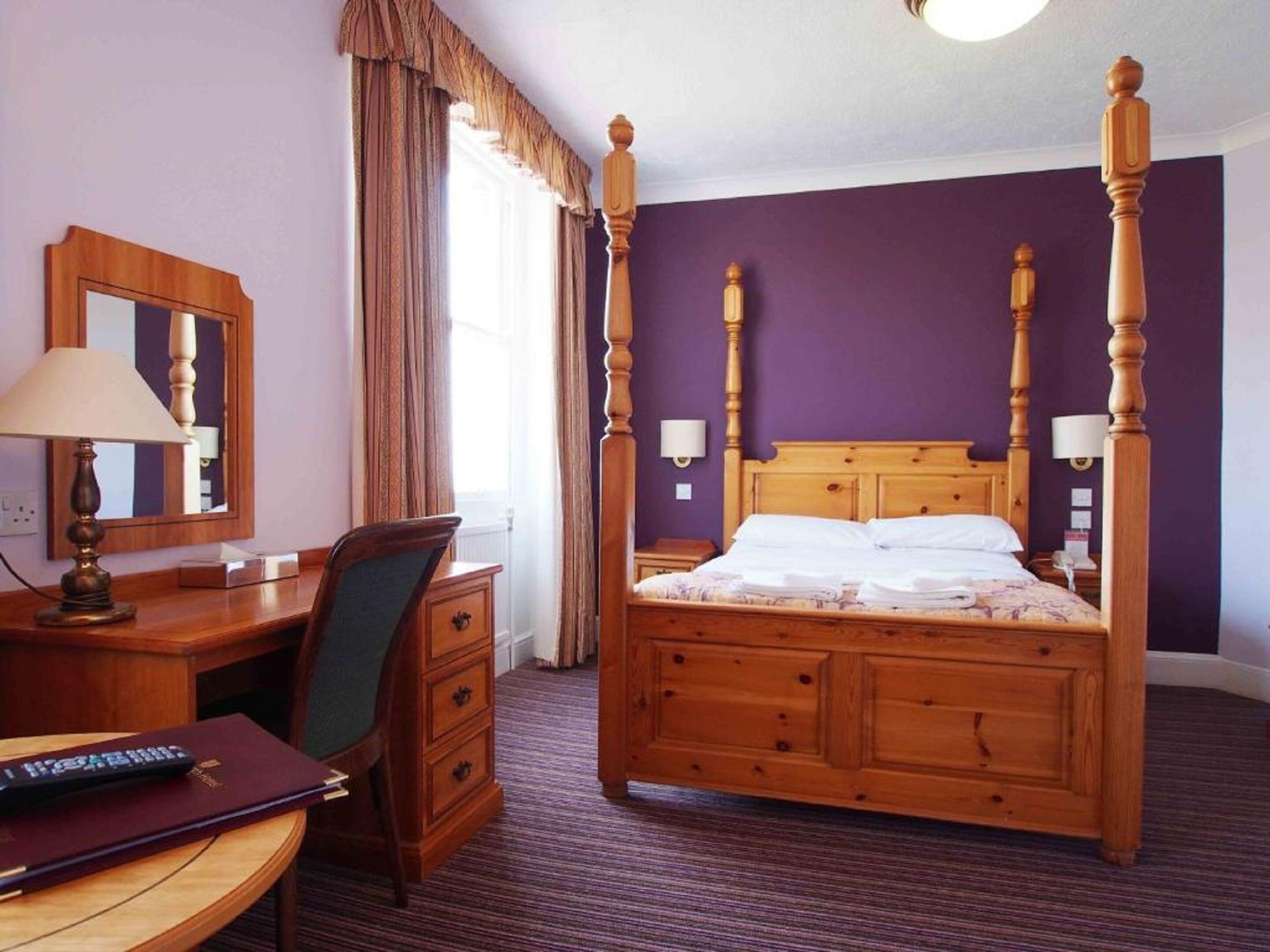 Un lit à baldaquin avec une tête de lit et un pied de lit en bois, un bureau avec une lampe, une chaise et une commode.