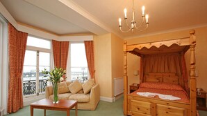 A bedroom with a four-poster bed, a wooden nightstand, a bedside table, a sofa, and a window with curtains. - Chatsworth Hotel - Worthing (Worthing)