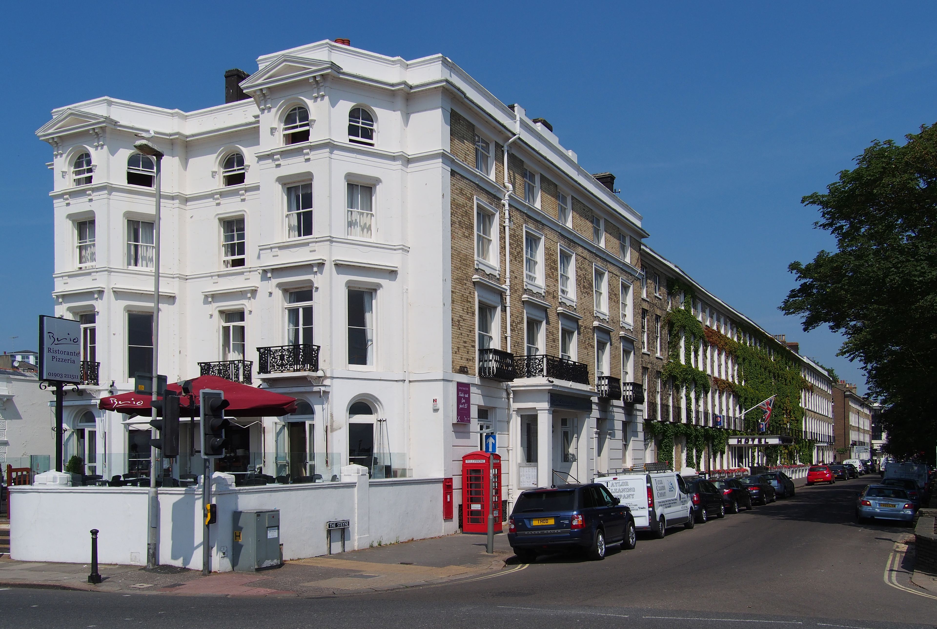 Un bâtiment blanc avec de nombreuses fenêtres et des balcons, une cabine téléphonique rouge et des voitures garées le long de la rue.