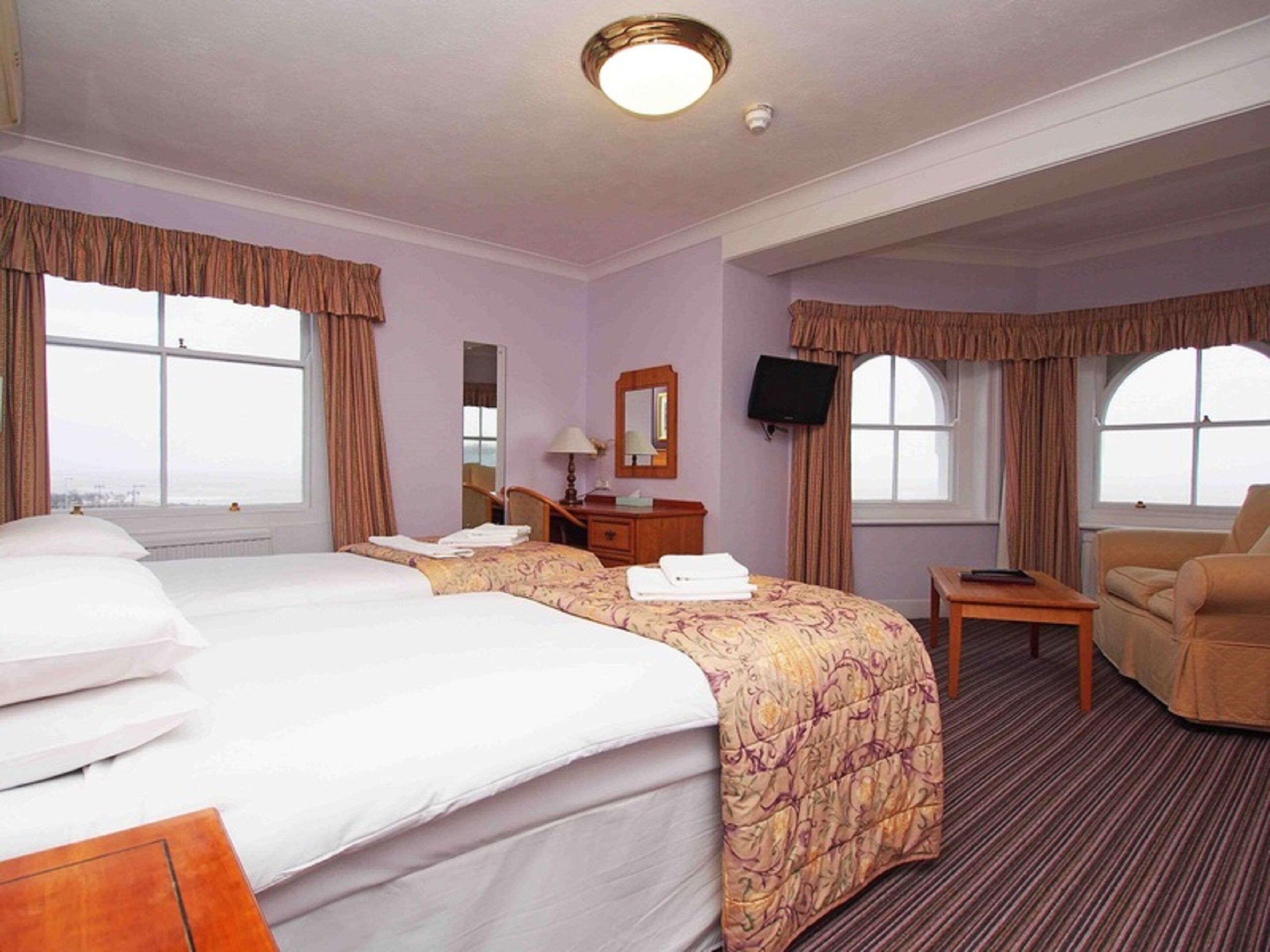 A hotel room with two beds, a desk, a chair, a television, and large windows.