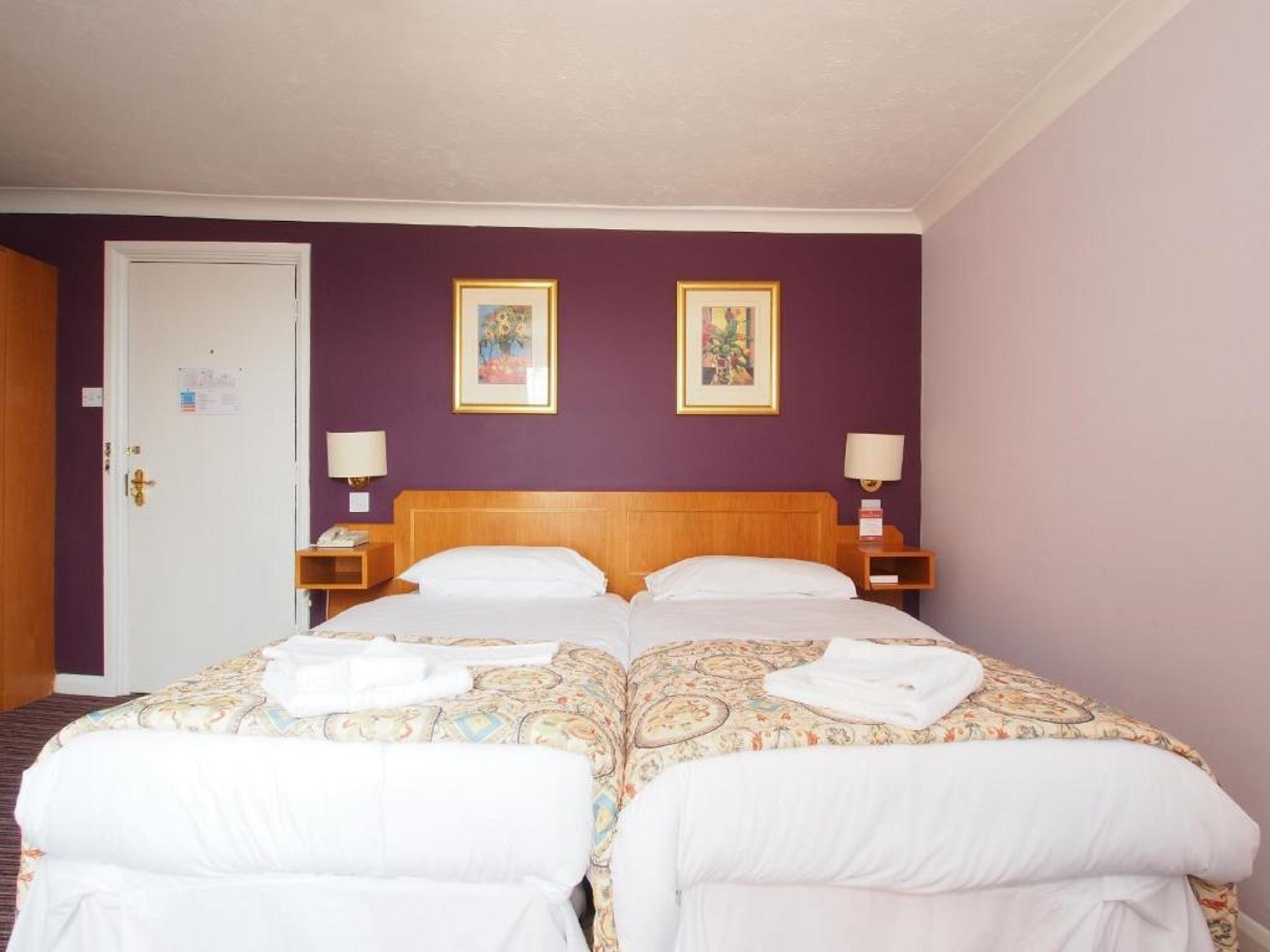 A hotel room with two beds, purple walls, and two framed pictures on the wall.