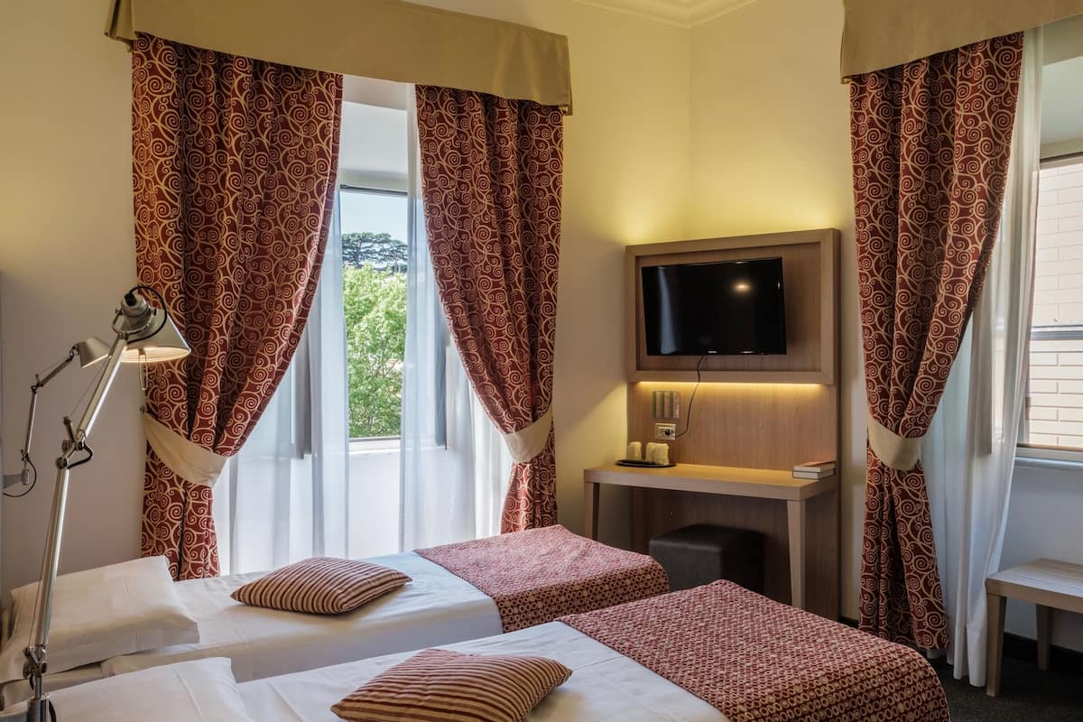 A hotel room with two beds, a TV mounted on the wall, a desk with a chair, and a window with curtains.