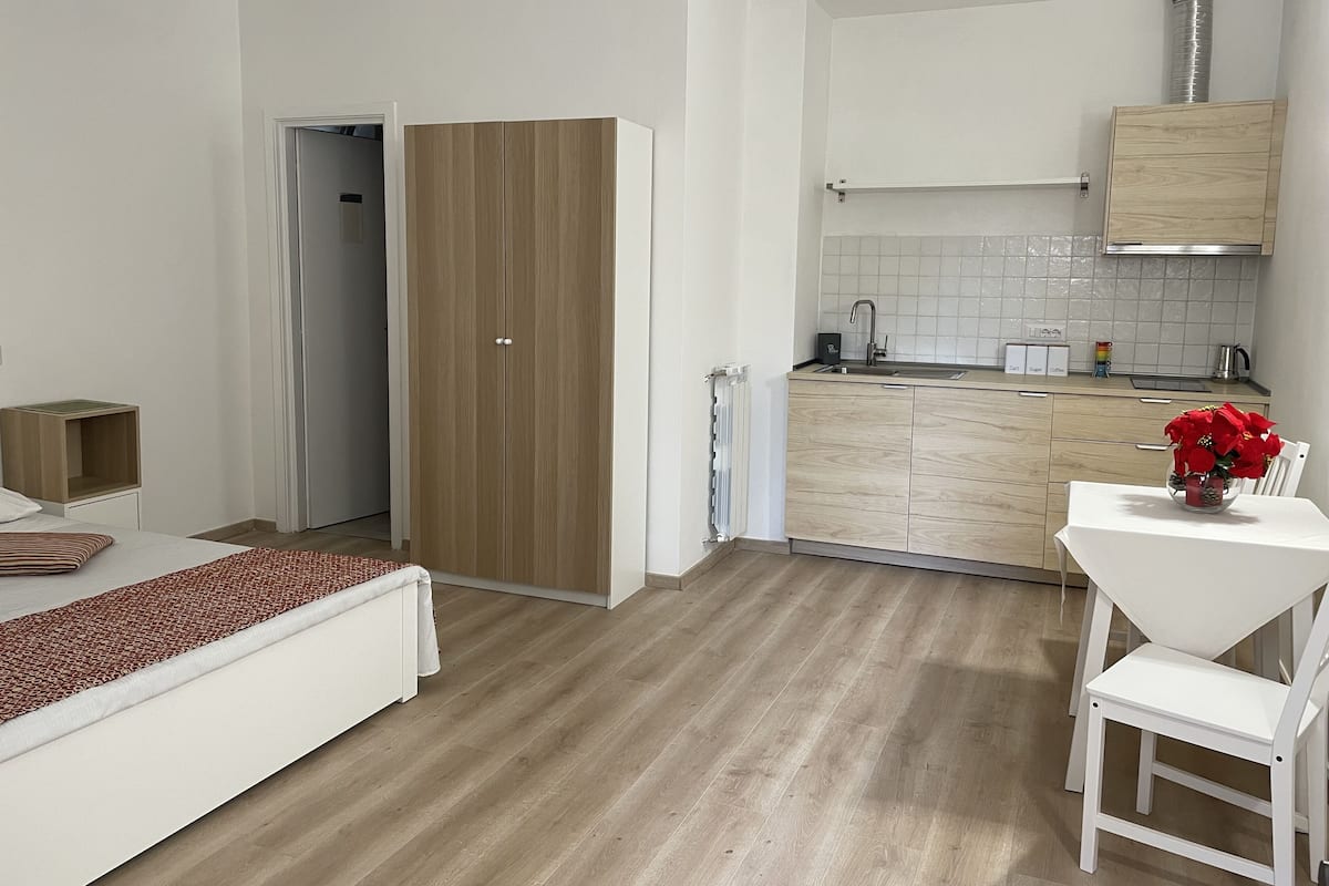 A modern, compact room with a wooden wardrobe, a small kitchenette, and a dining area with a white table and chair.