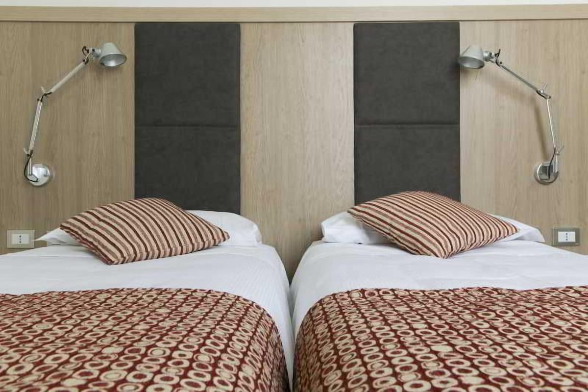 A hotel room with two beds, each with a patterned bedspread and striped pillows. There are two wall-mounted lamps on either side of the beds.