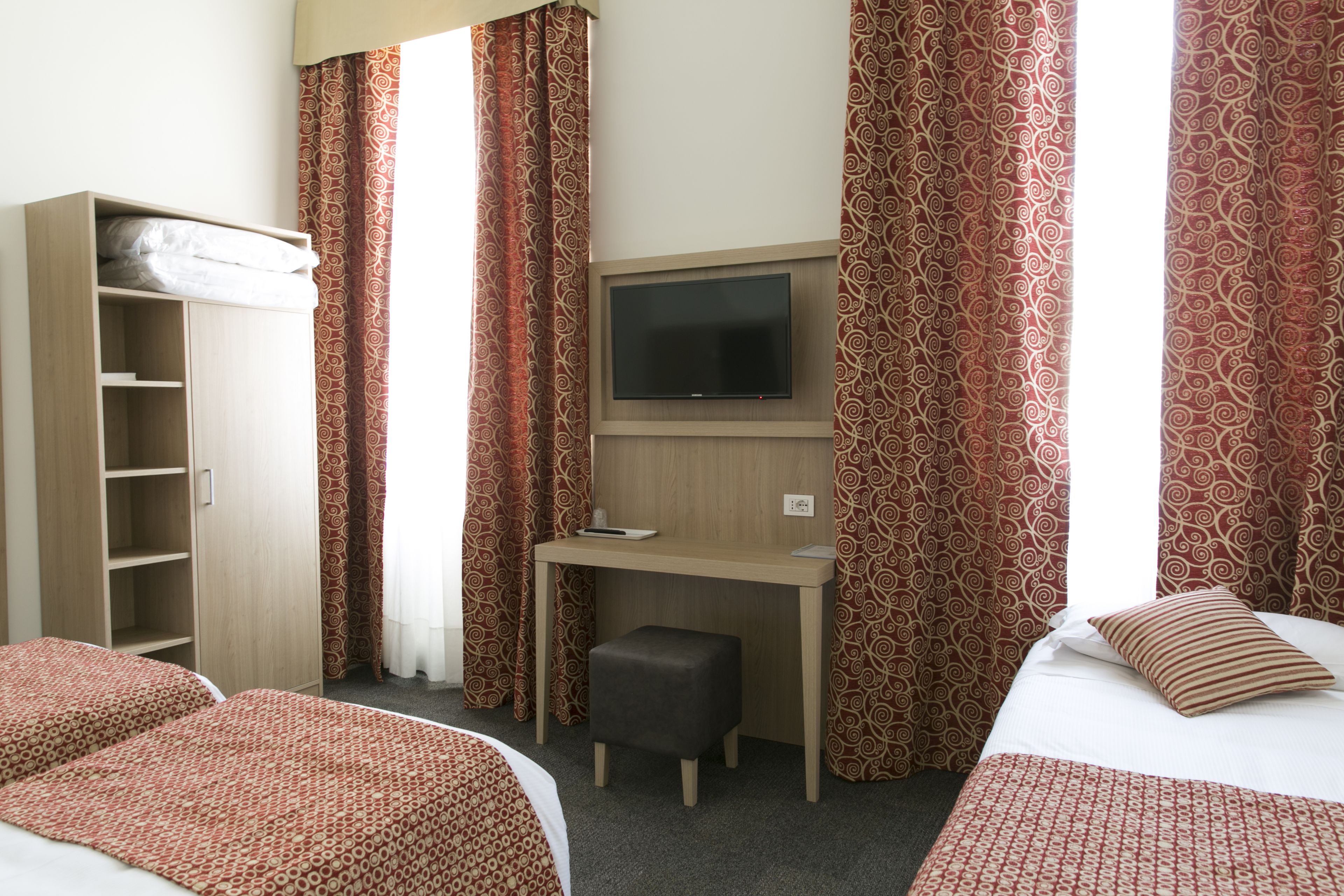 A hotel room with two beds, a desk with a TV, a wardrobe, and patterned curtains.