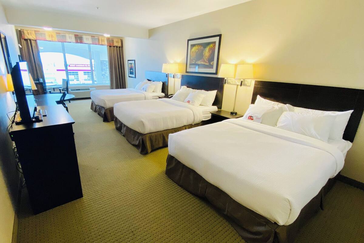A hotel room with two beds, a desk, and a chair. There is a window with curtains, a painting on the wall, and a lamp on the desk.