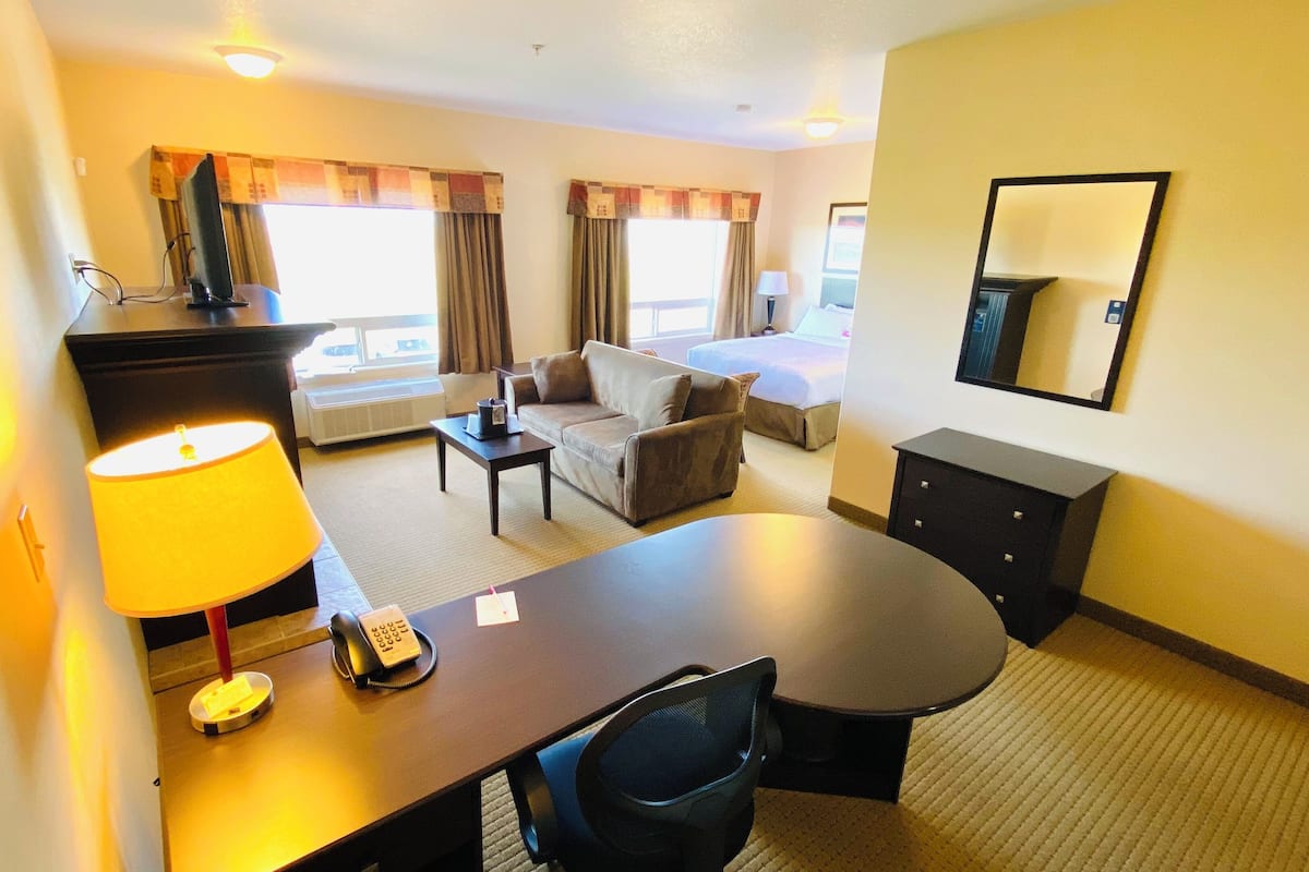 A hotel room with a dining table, a sofa, a chair, a desk with a lamp, a telephone, and a bed.