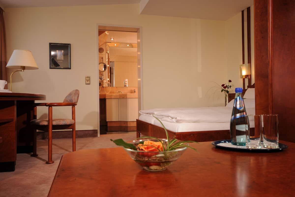 A hotel room with a bed, a desk with a lamp, a chair, a bedside table with a vase and glasses, and a bathroom visible through an open door.