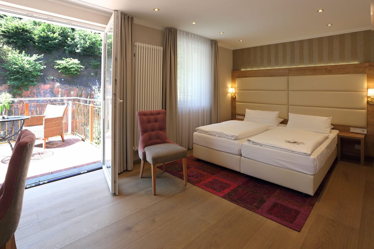A hotel room with a double bed, a chair, a small table, and a view of greenery through the window.