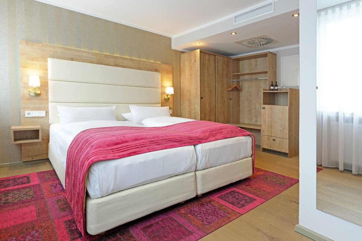 A modern hotel room with a large bed, wooden wardrobe, and a small seating area.