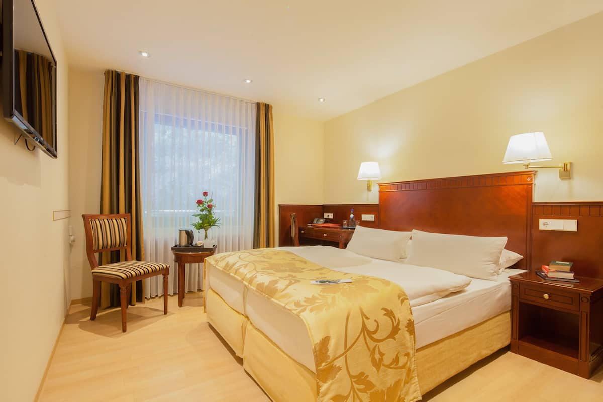 A hotel room with a double bed, a bedside table with a lamp, a chair, a small table with a vase of flowers, and a television mounted on the wall.