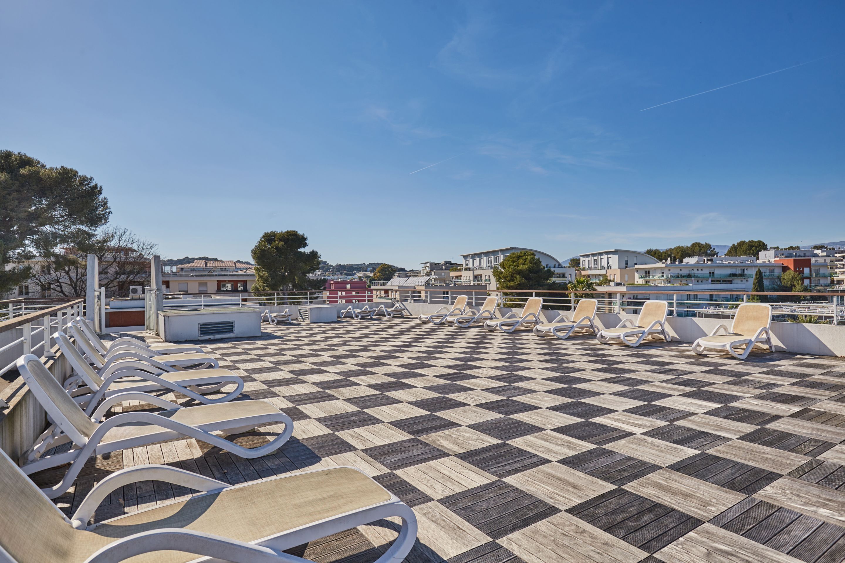A checkered deck with lounge chairs, a railing, and buildings in the background.