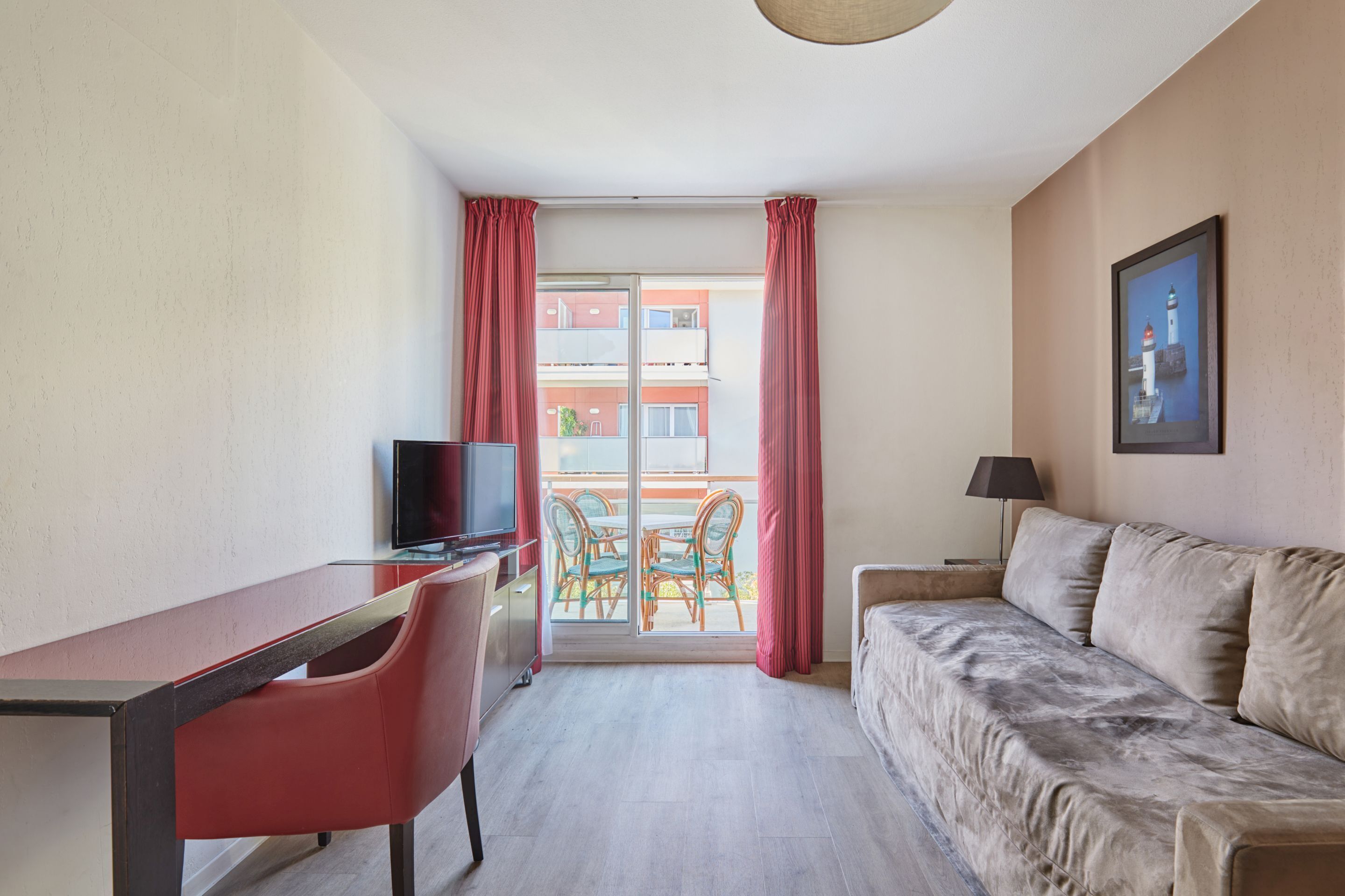 A room with a bed, a desk with a chair, a television, a table with chairs, and a balcony with red curtains.
