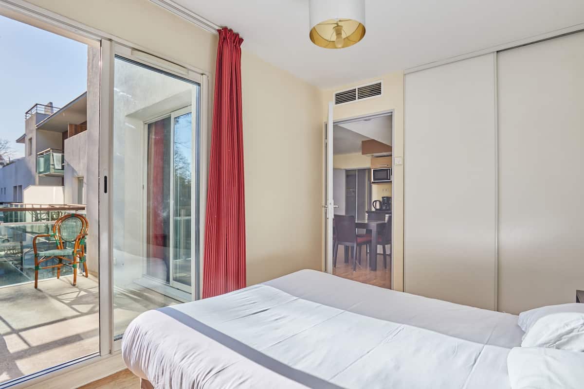 A bedroom with a bed, a sliding glass door to a balcony, a dining area, and a kitchenette.