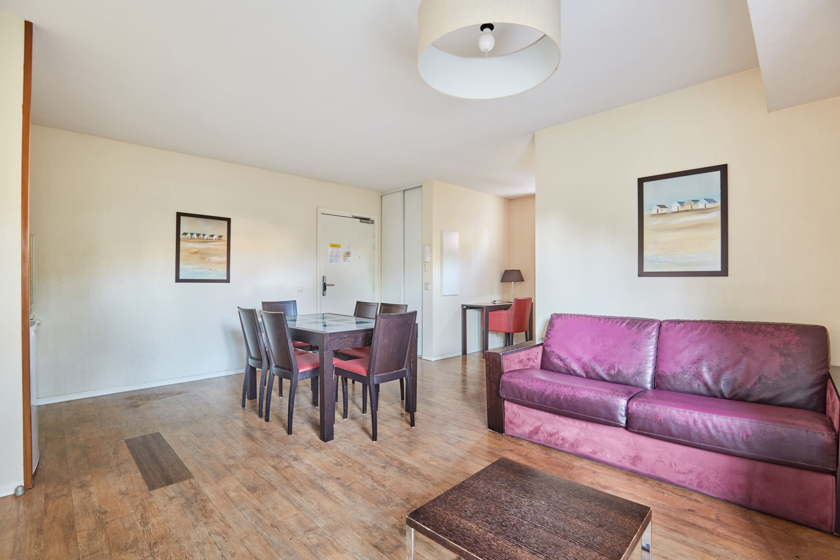 Een woonkamer met houten vloer, een paarse bank, een glazen eettafel met stoelen en een plafondlamp.