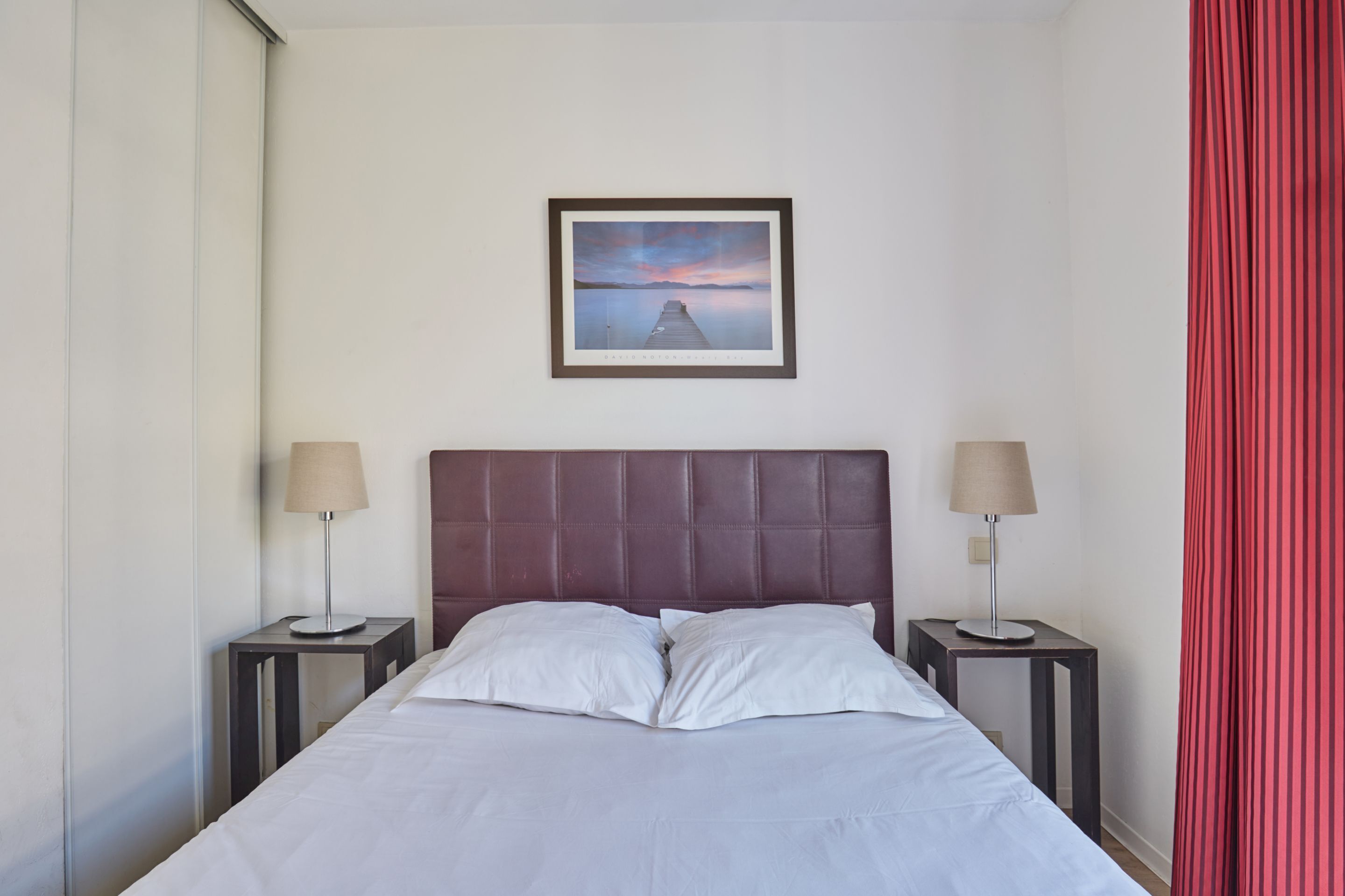 Habitación de hotel con cama, dos mesitas de noche, cabecera y un cuadro enmarcado en la pared, además de una cortina roja.