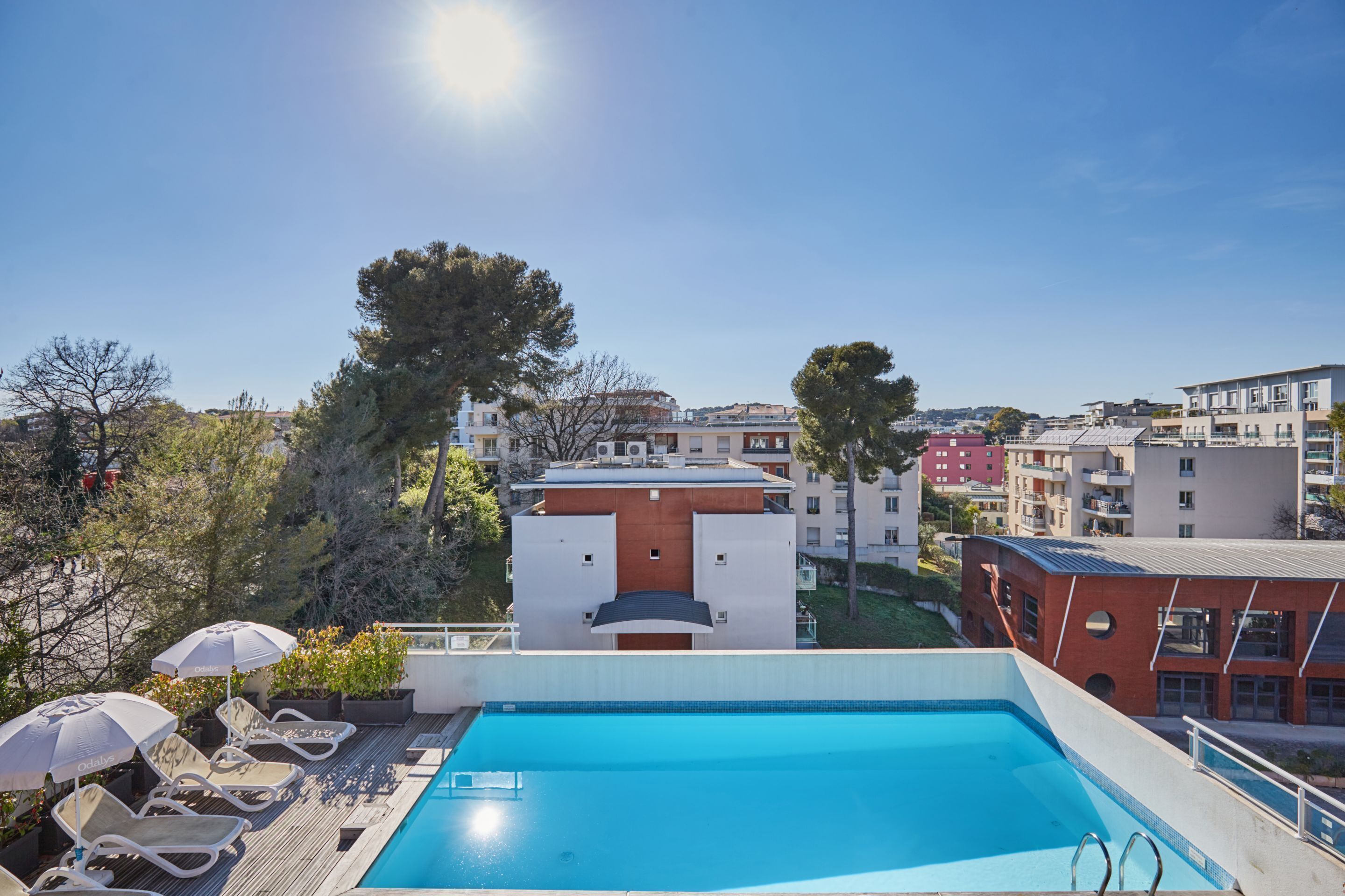 A rooftop pool with lounge chairs and umbrellas, surrounded by buildings and trees.