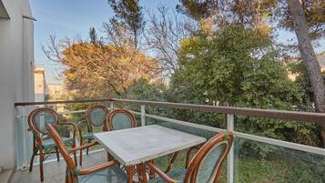 Un balcone con sedie in vimini e un tavolo, con vista su un giardino alberato e un edificio sullo sfondo.