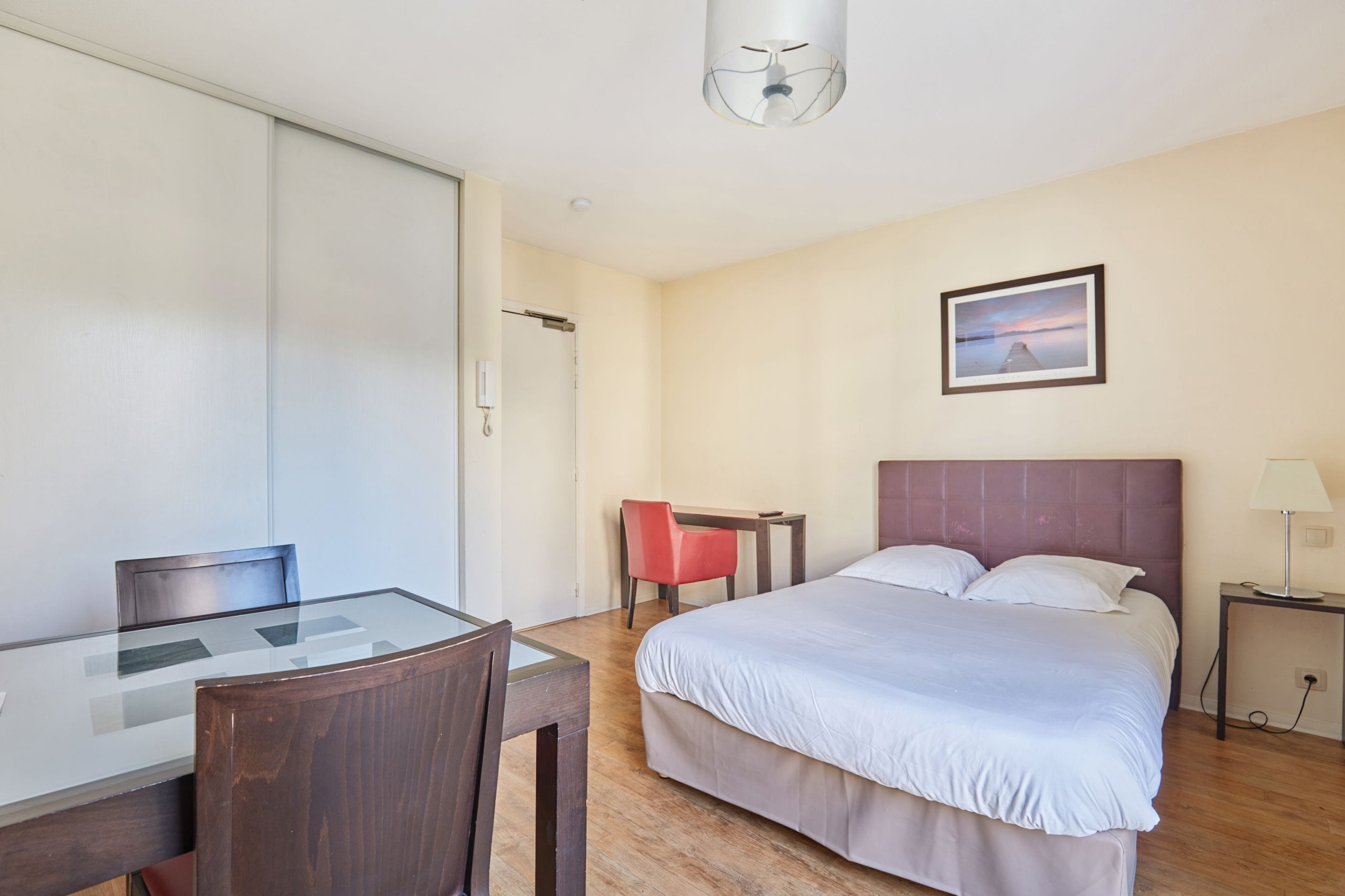 A hotel room with a bed, a desk with a chair, a small table, a lamp, and a picture on the wall.