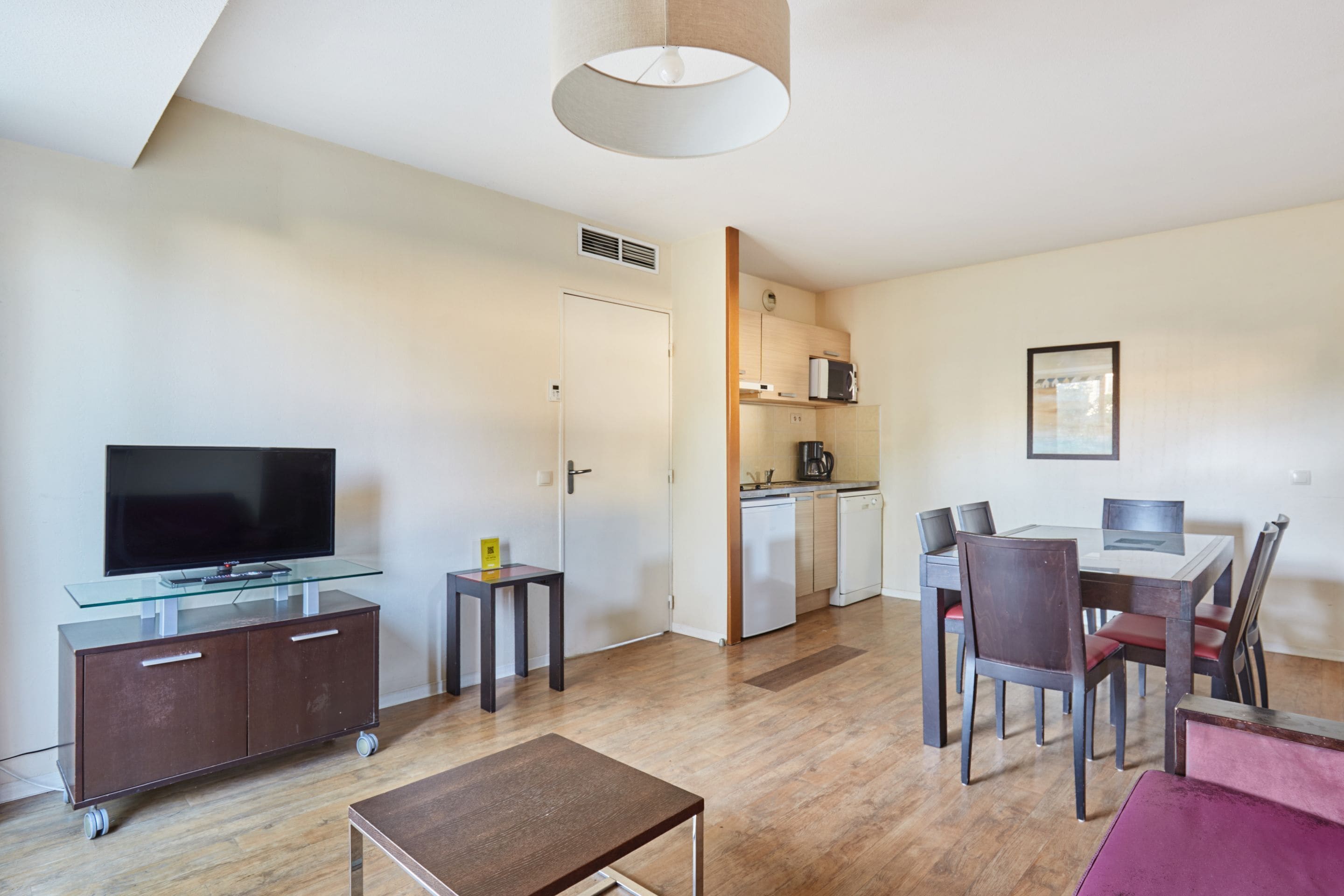 A modern living room with a flat-screen TV, a glass-top coffee table, a dining area with a table and chairs, a kitchen with a dishwasher, and a ceiling light fixture.