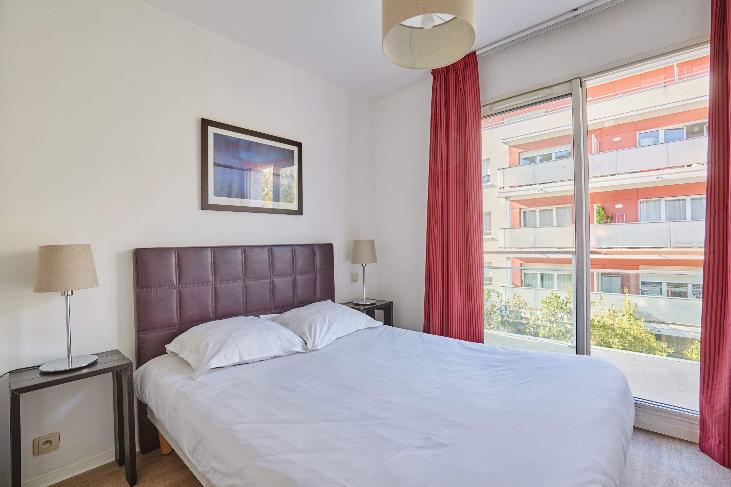 Une chambre d’hôtel avec un grand lit, deux tables de chevet avec des lampes, un tableau encadré au mur et une vue sur un immeuble à travers une fenêtre ornée de rideaux rouges.