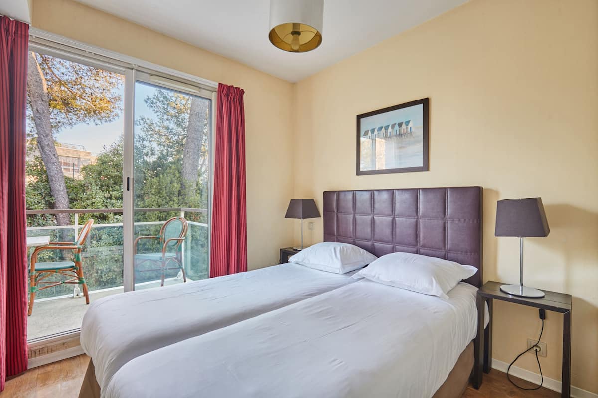 Ein Hotelzimmer mit einem großen Bett, zwei Nachttischen, einem Fenster mit Vorhängen und einem Balkon mit Sitzgelegenheiten.